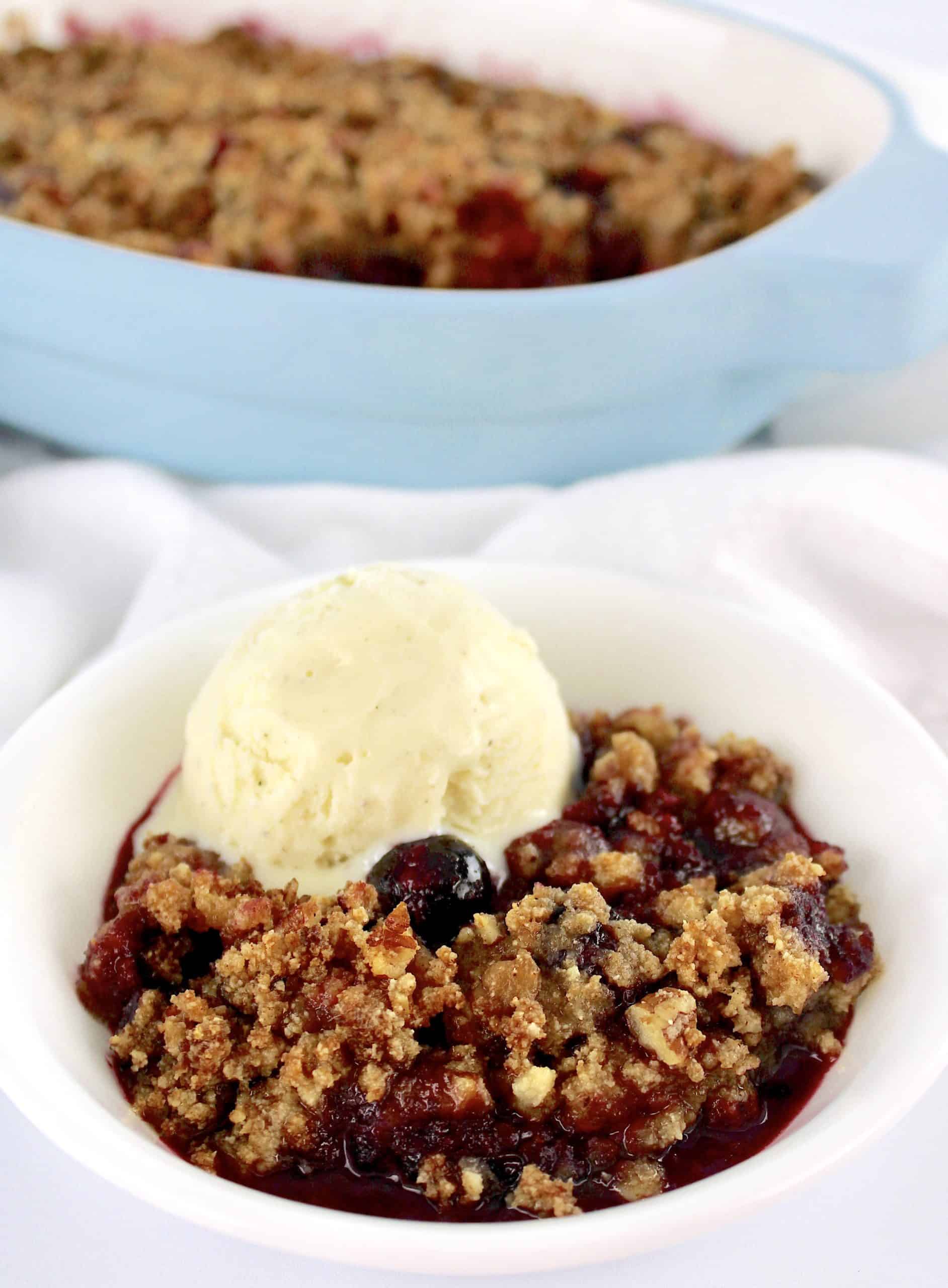 Mixed Berry Crumble in white bowl with scoop of vanilla ice cream and casserole in background