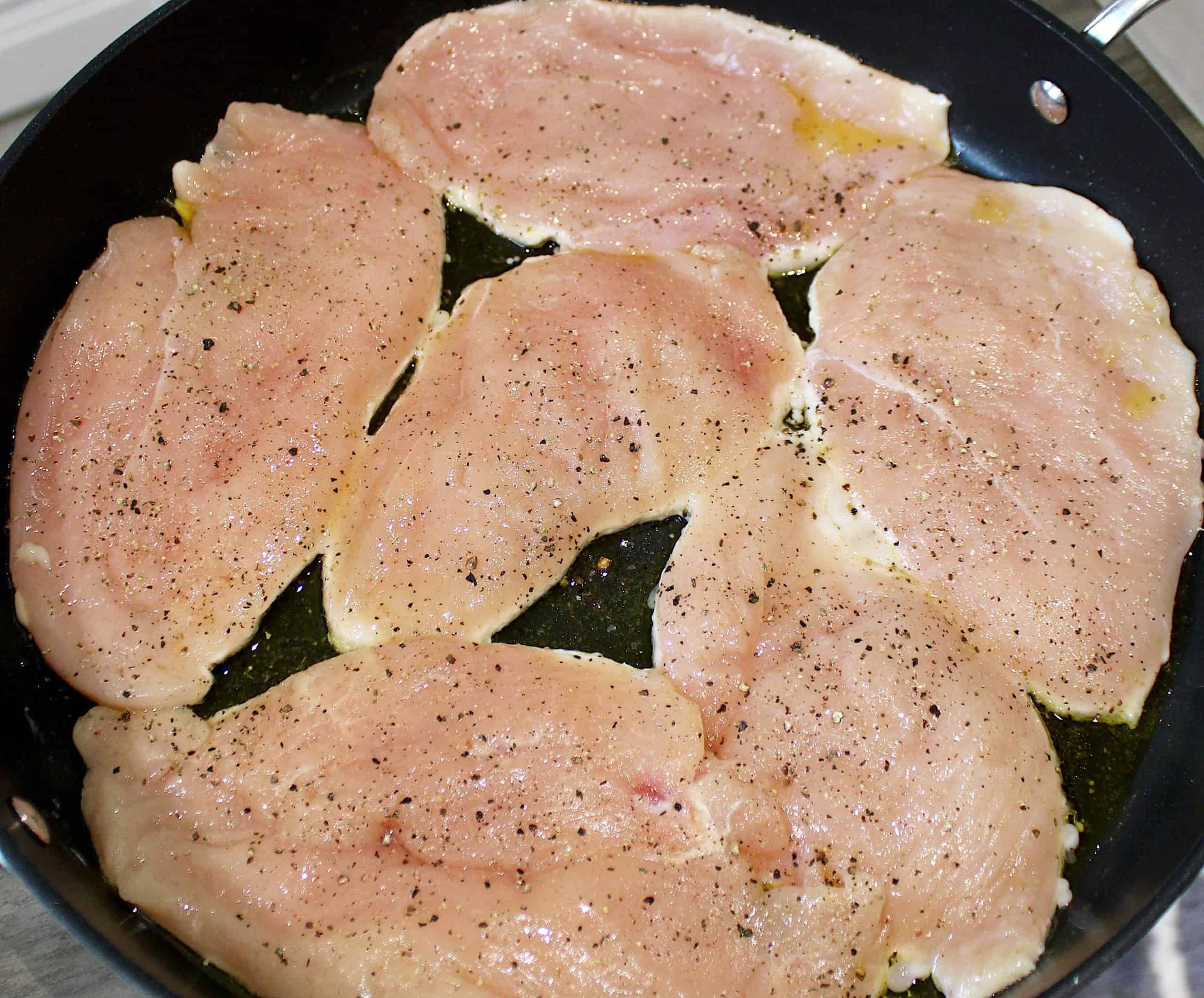 6 chicken breasts cooking in skillet with salt and pepper on top