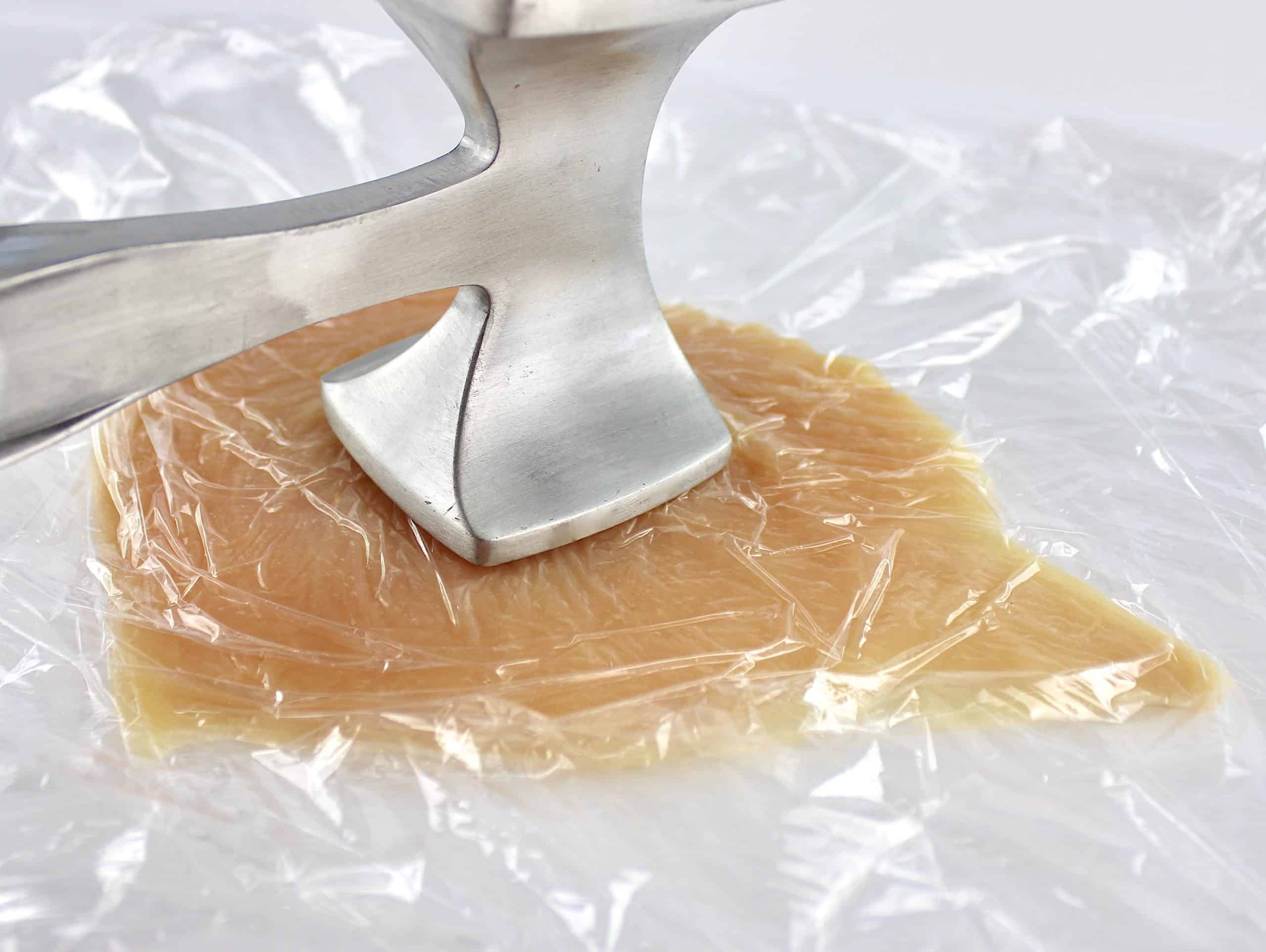 chicken breast in plastic wrap being pounded with silver mallet