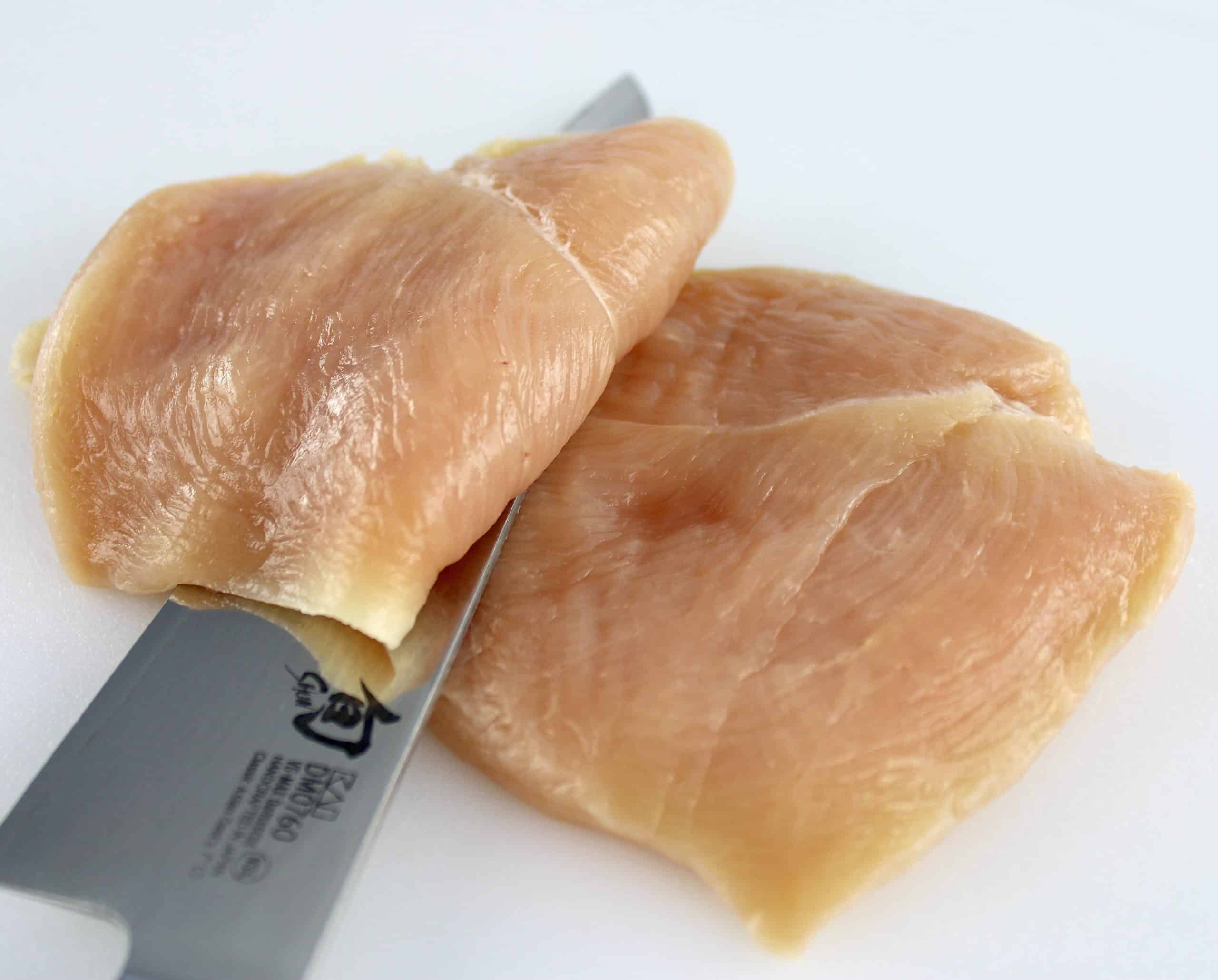 chicken breast being sliced in half on white cutting board