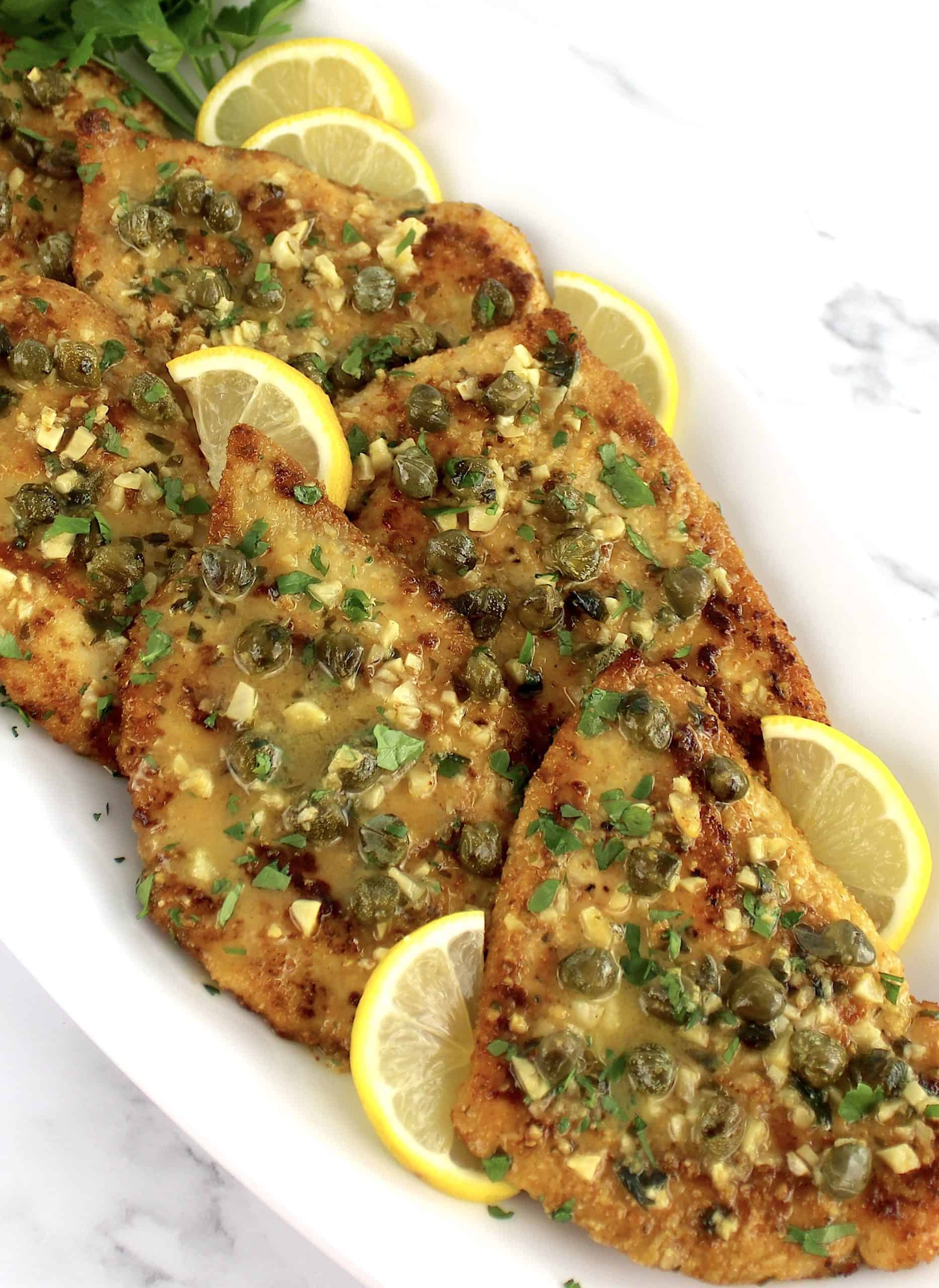 Chicken Piccata on white platter with lemon slices and chopped parsley