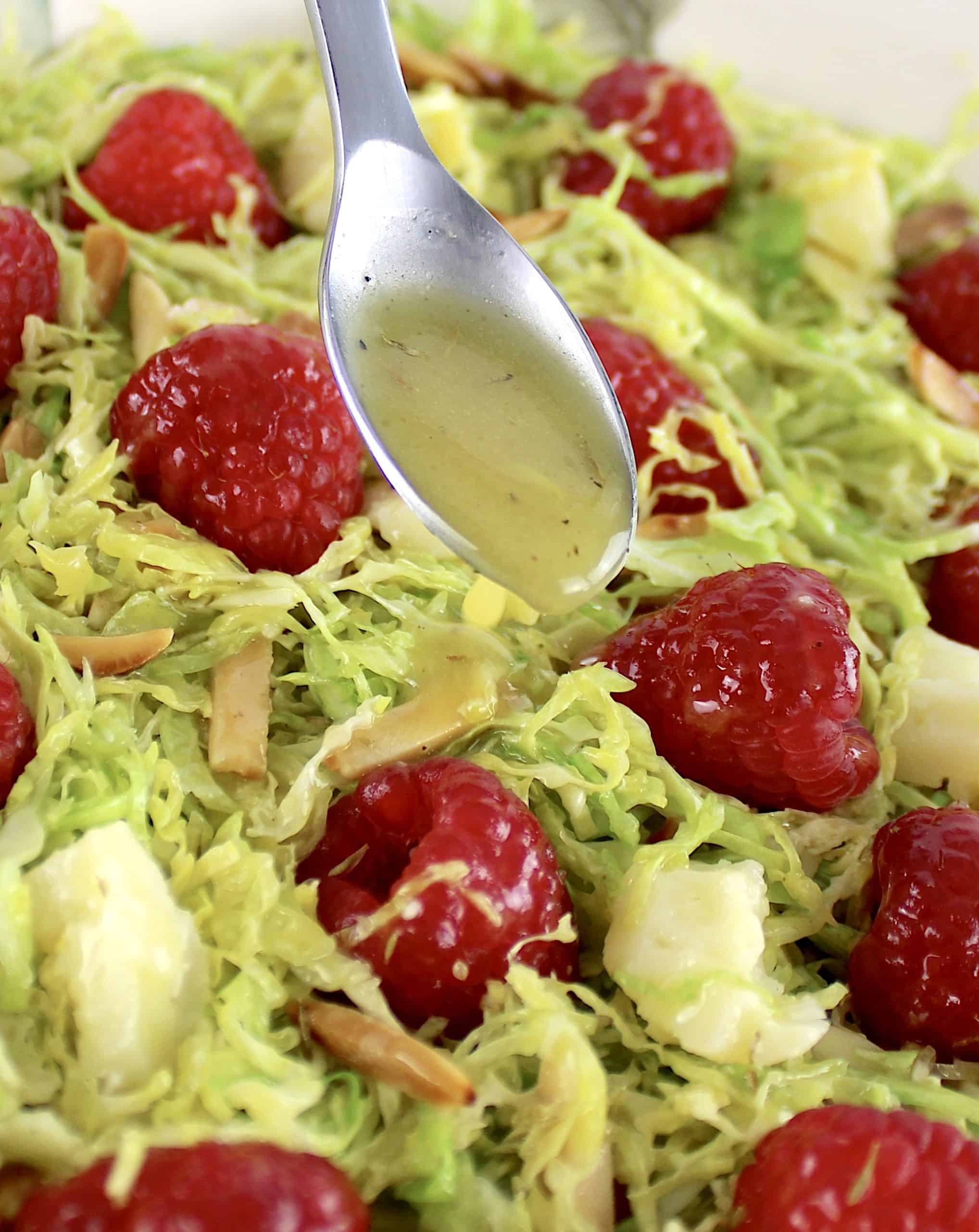 Shaved Brussels Sprouts Salad with raspberries parmesan cheese and slivered almonds with dressing being spooned over top