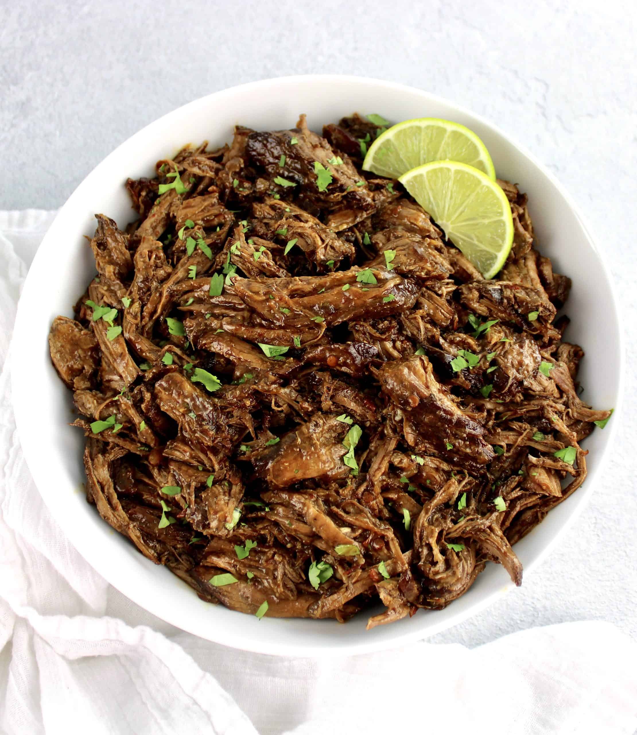 Slow Cooker Beef Barbacoa in white bowl with chopped cilantro and lime slices