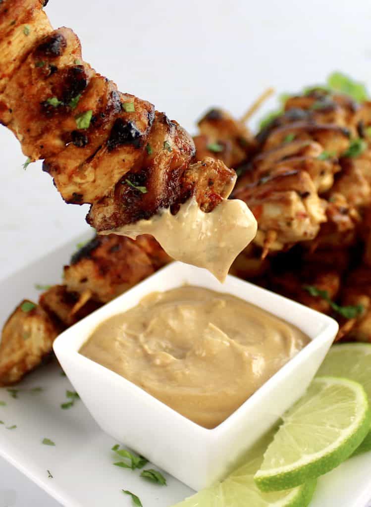 thai chicken satay dipping into sauce on white plate with lime slices on side