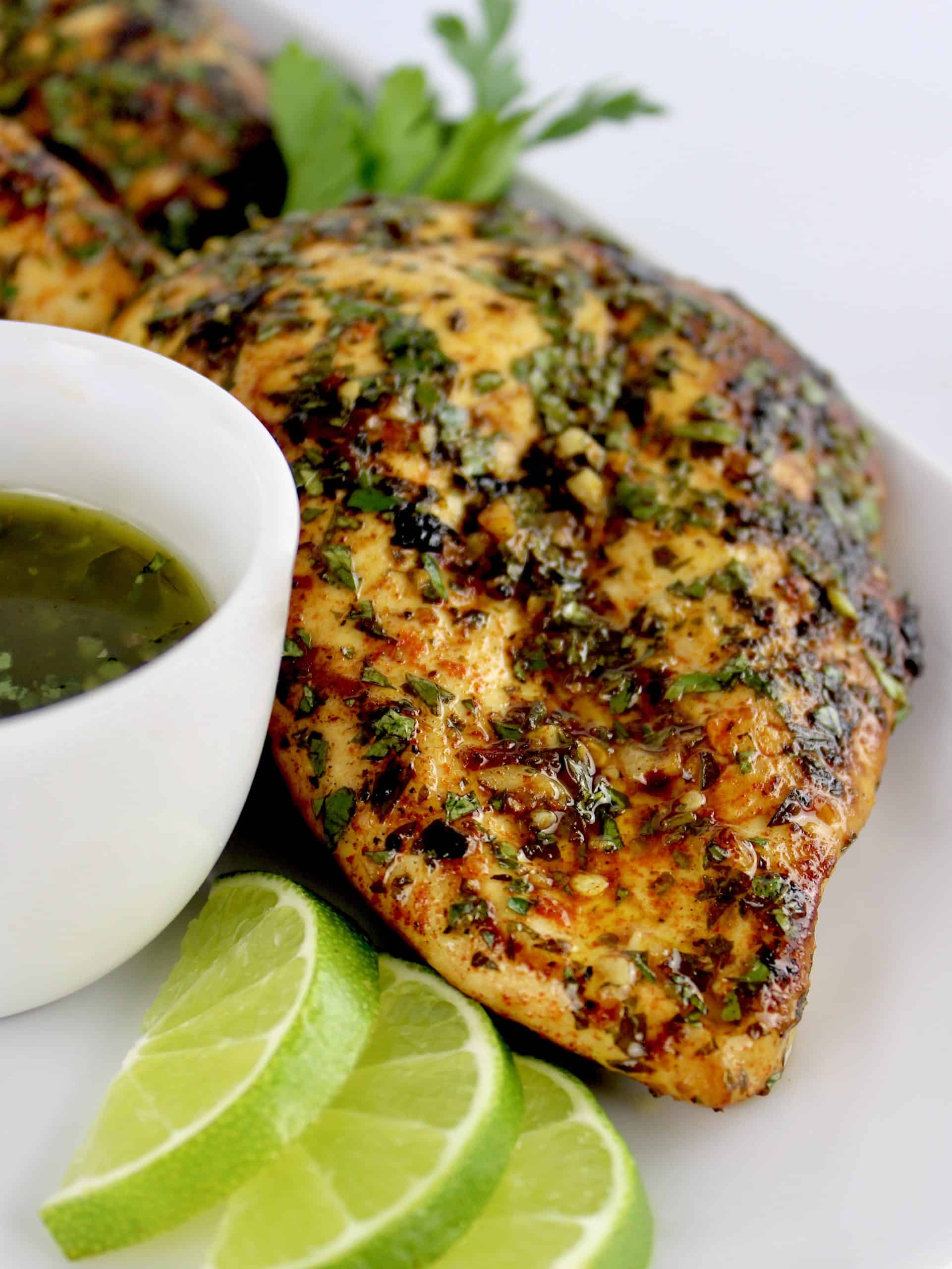 closeup of Chimichurri chicken on white plate with sauce in white cup on side and lime slices
