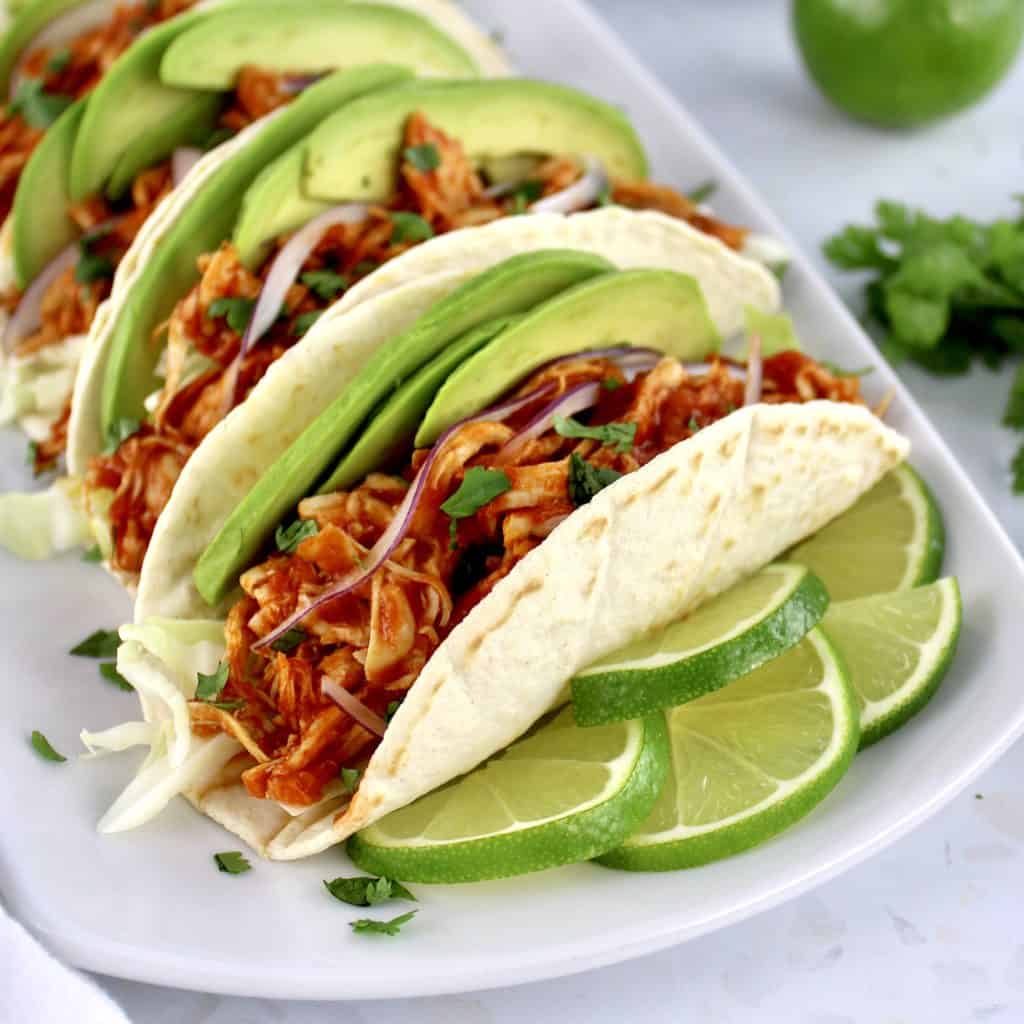 closeup of Chicken Tinga in tortillas on white plate with limes slices on side