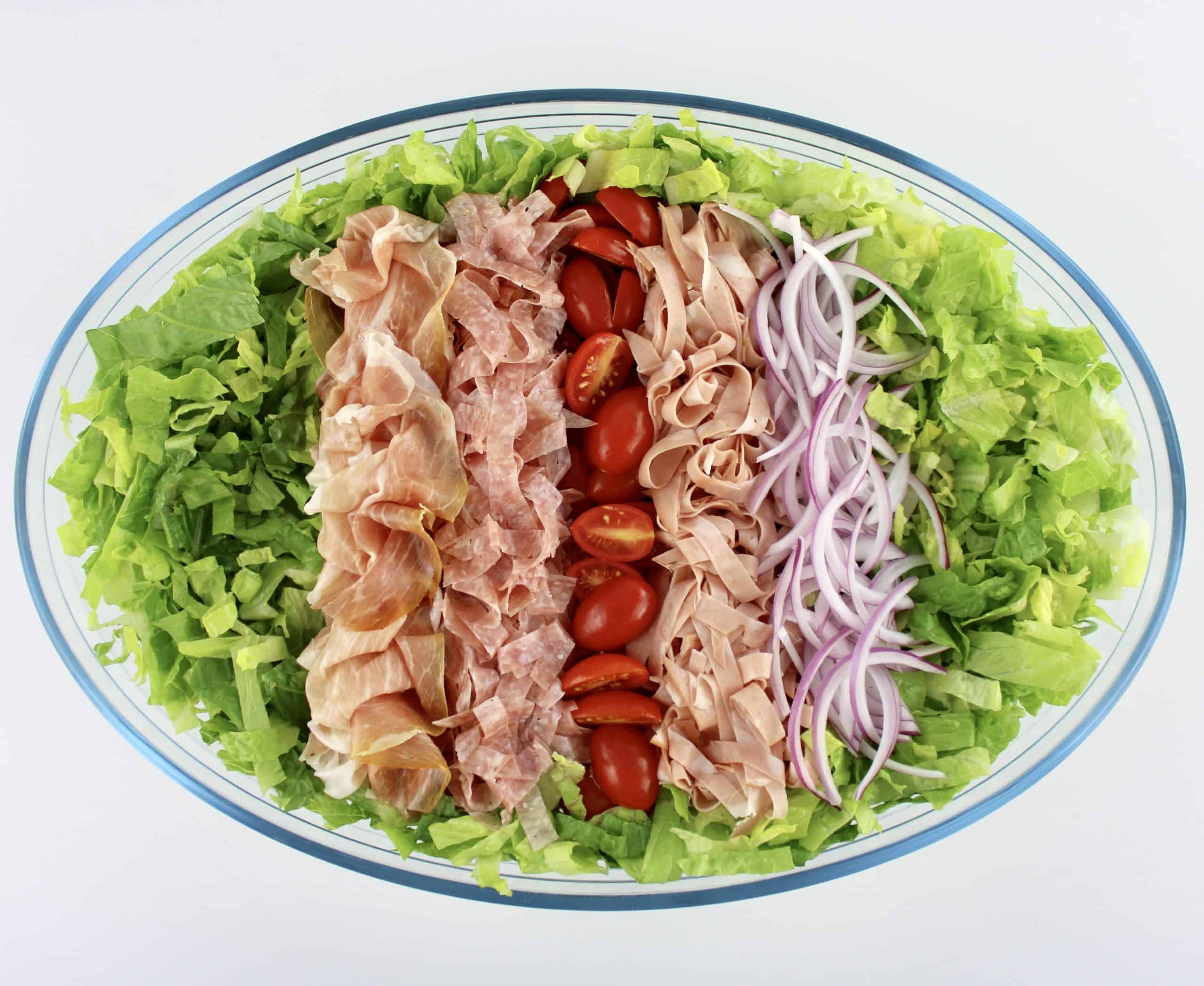 shredded lettuce in oval glass bowl with rows of tomatoes and shredded salami and mortadella in the center with prosciutto and sliced red onions on the sides