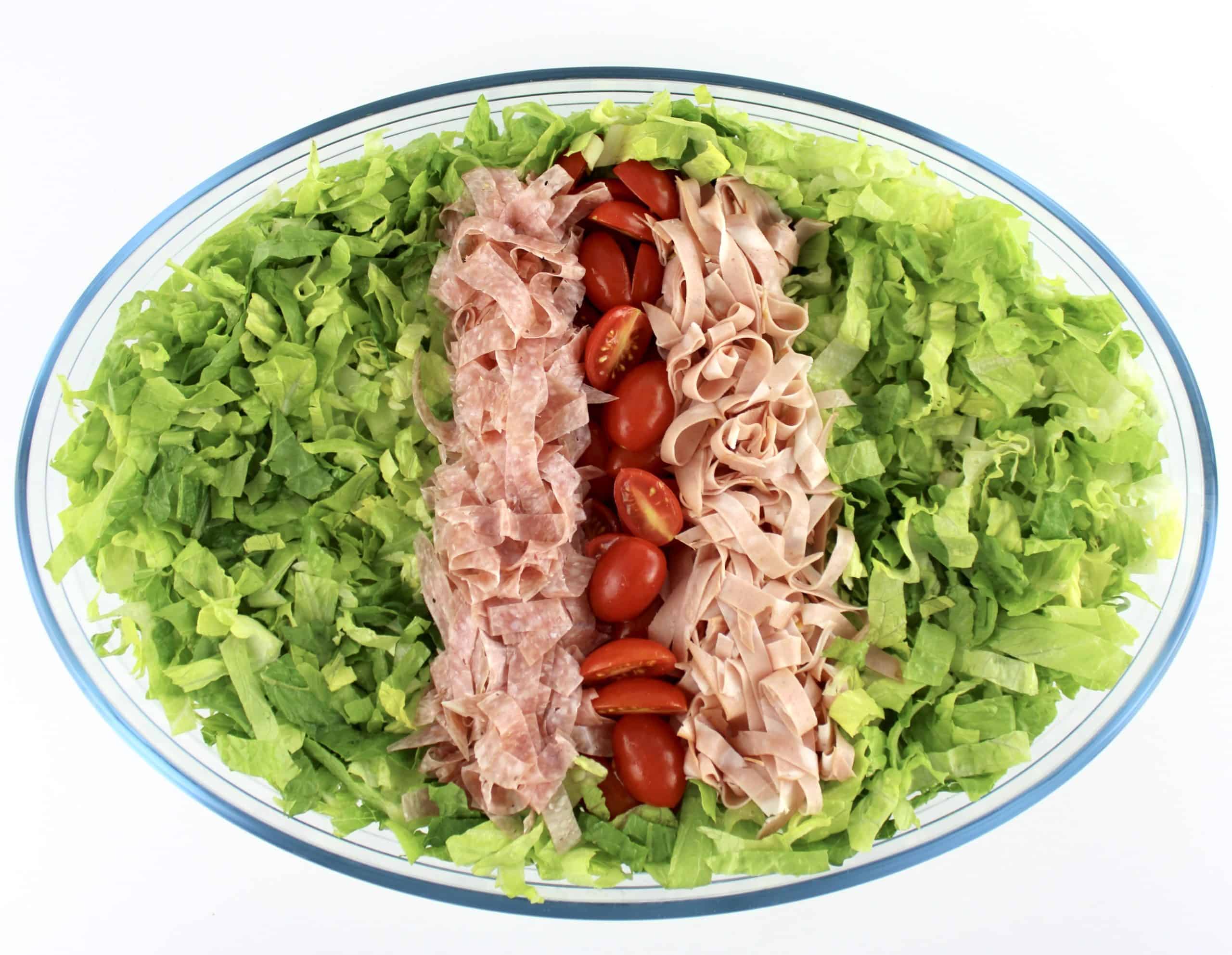 shredded lettuce in oval glass bowl with rows of tomatoes and shredded salami and mortadella in the center