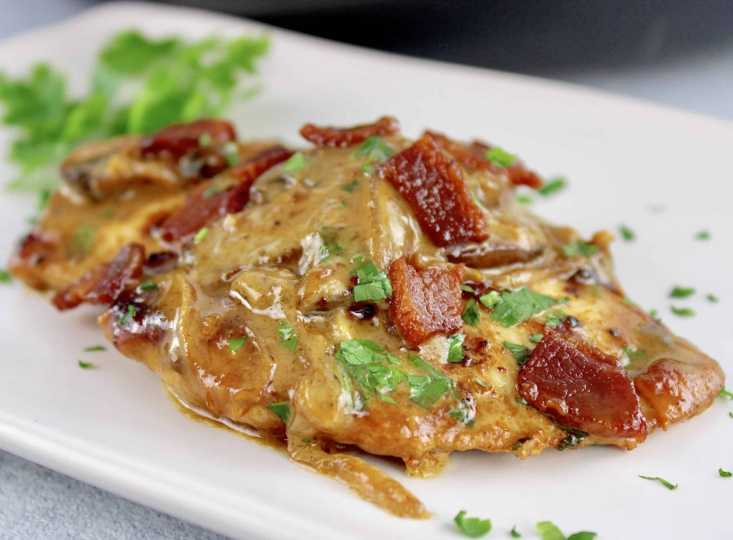 closeup of piece of Creamy Bacon Mushroom Chicken on white plate with chopped parsley and bacon on top