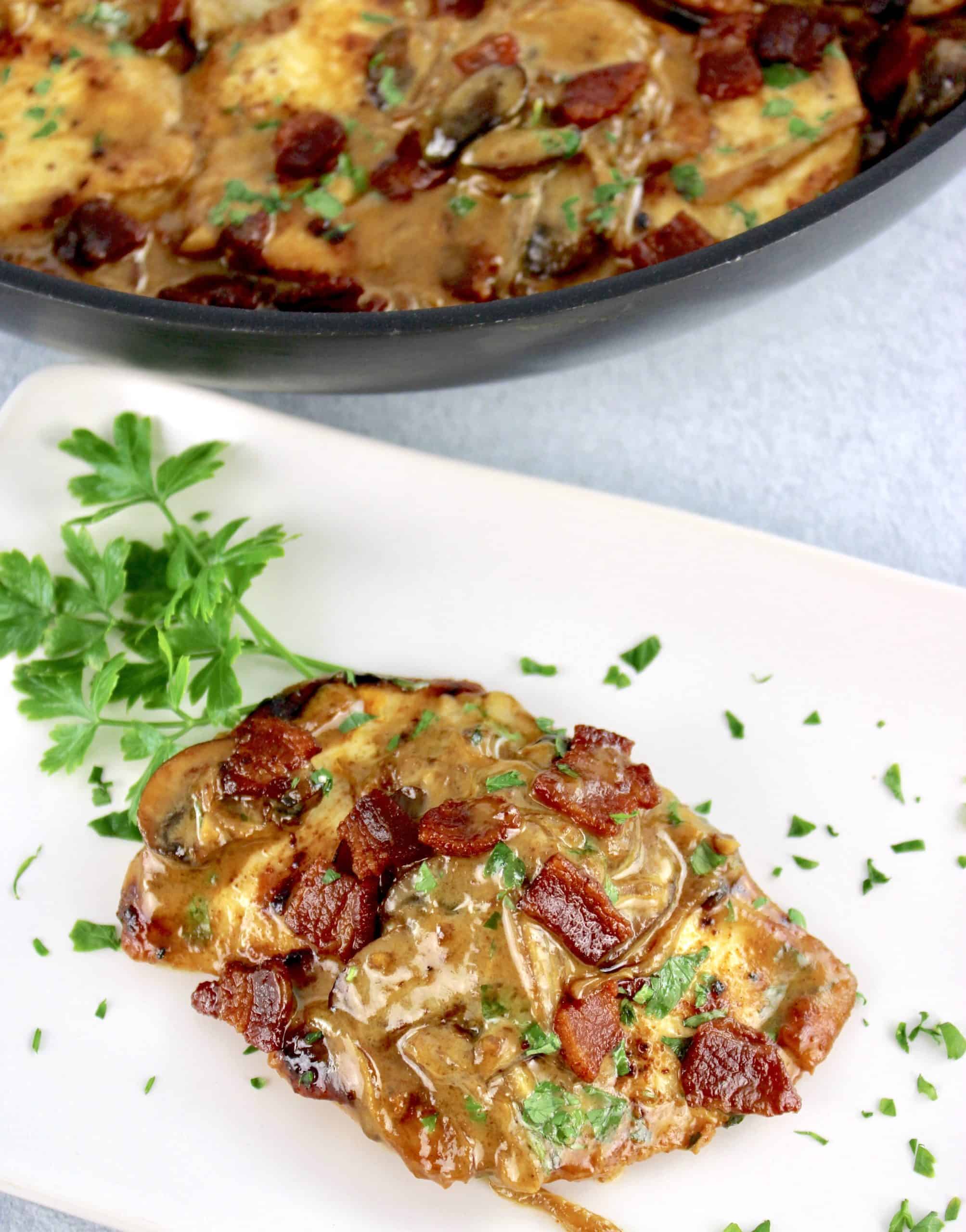 Creamy Bacon Mushroom Chicken piece on white plate with parsley garnish and skillet in background