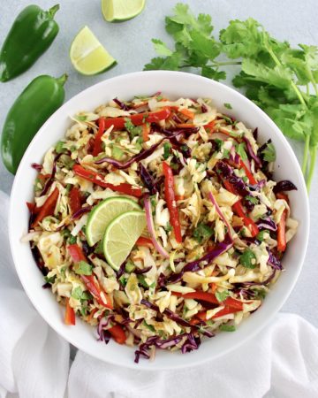 overhead view of Mexican Coleslaw in white bowl with lime cilantro and jalapeños in background