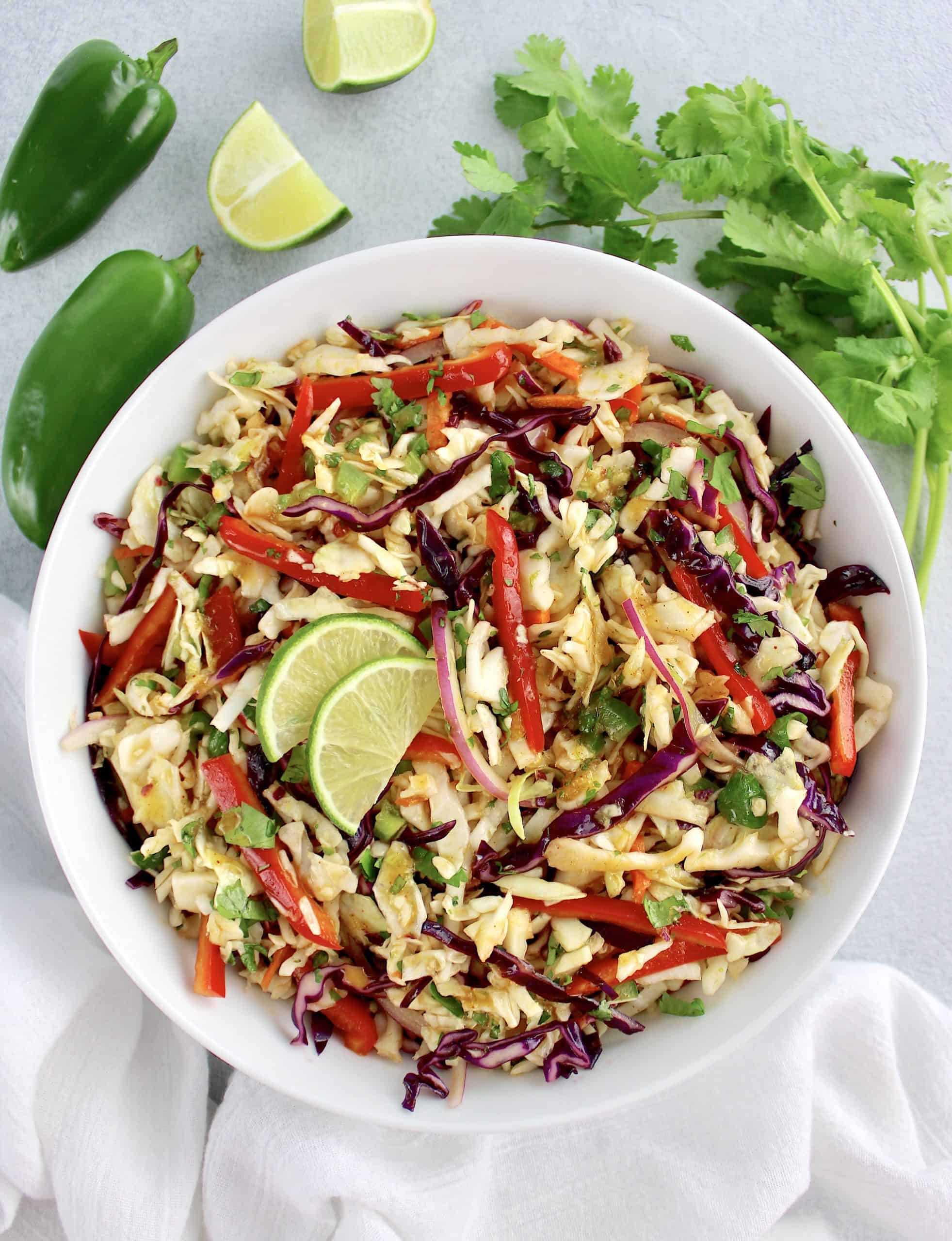 overhead view of Mexican Coleslaw in white bowl with lime cilantro and jalapeños in background