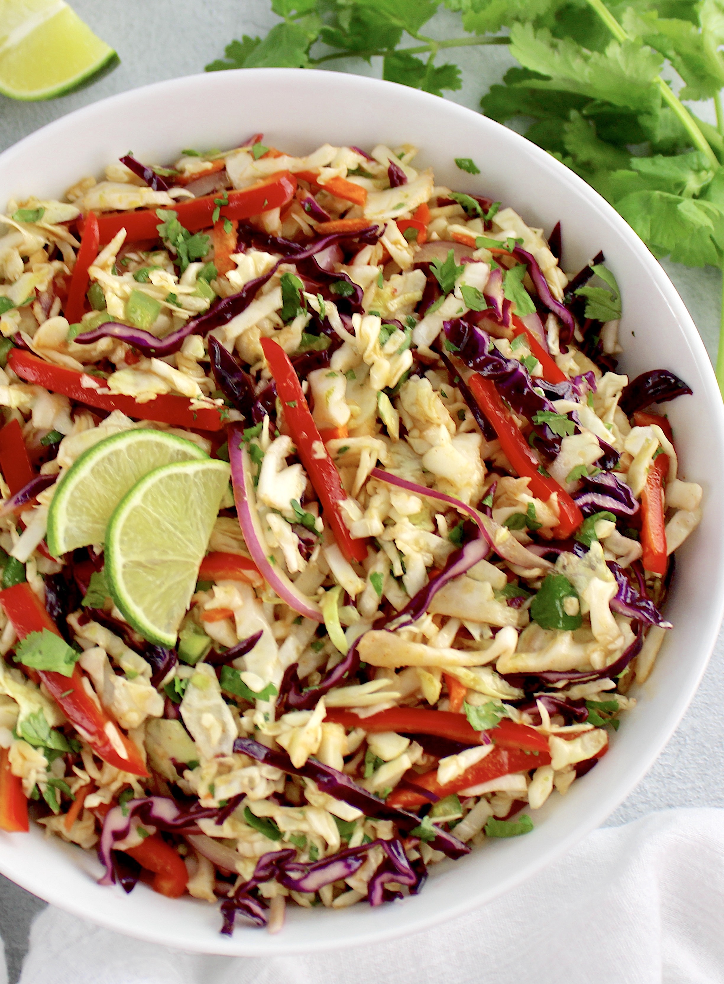 overhead view of Mexican Coleslaw in white bowl with lime slices ad cilantro in background