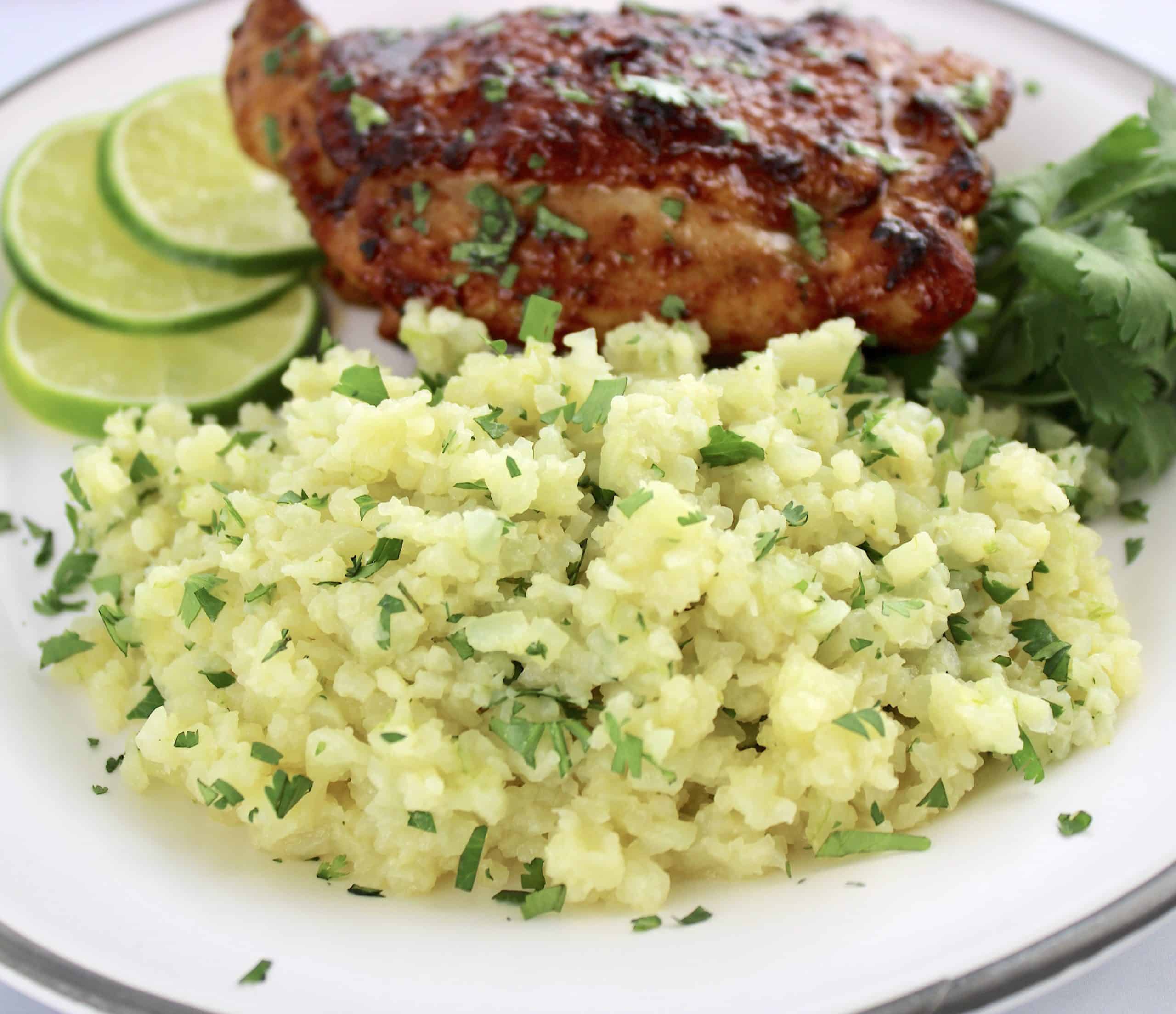 closeup of lime cilantro cauliflower rice on white plate with chicken thigh and lime slices in back