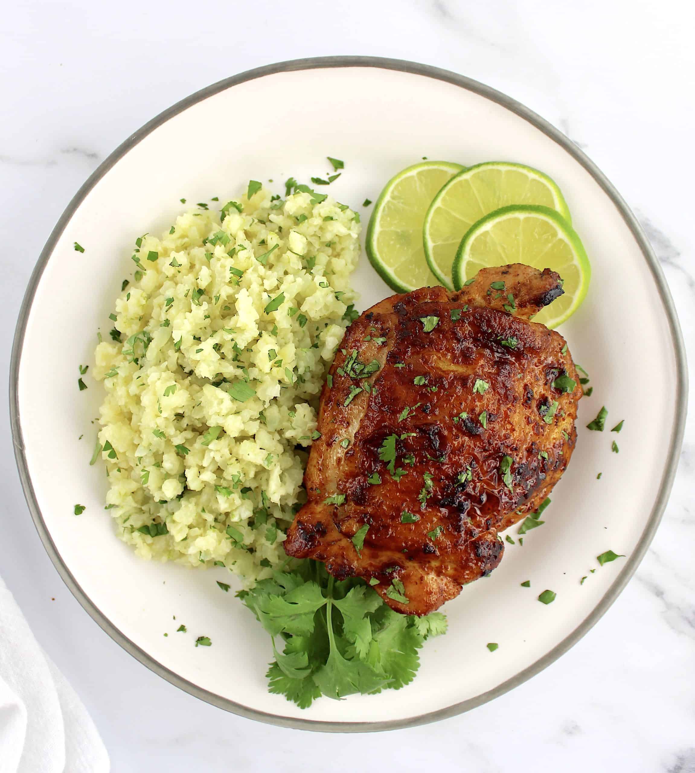 Paprika Chicken Thigh on white plate with cilantro cauliflower rice cilantro and lime slices on side