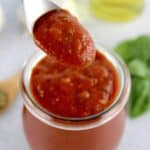 pizza sauce being spooned out of open jar with basil and olive oil in background
