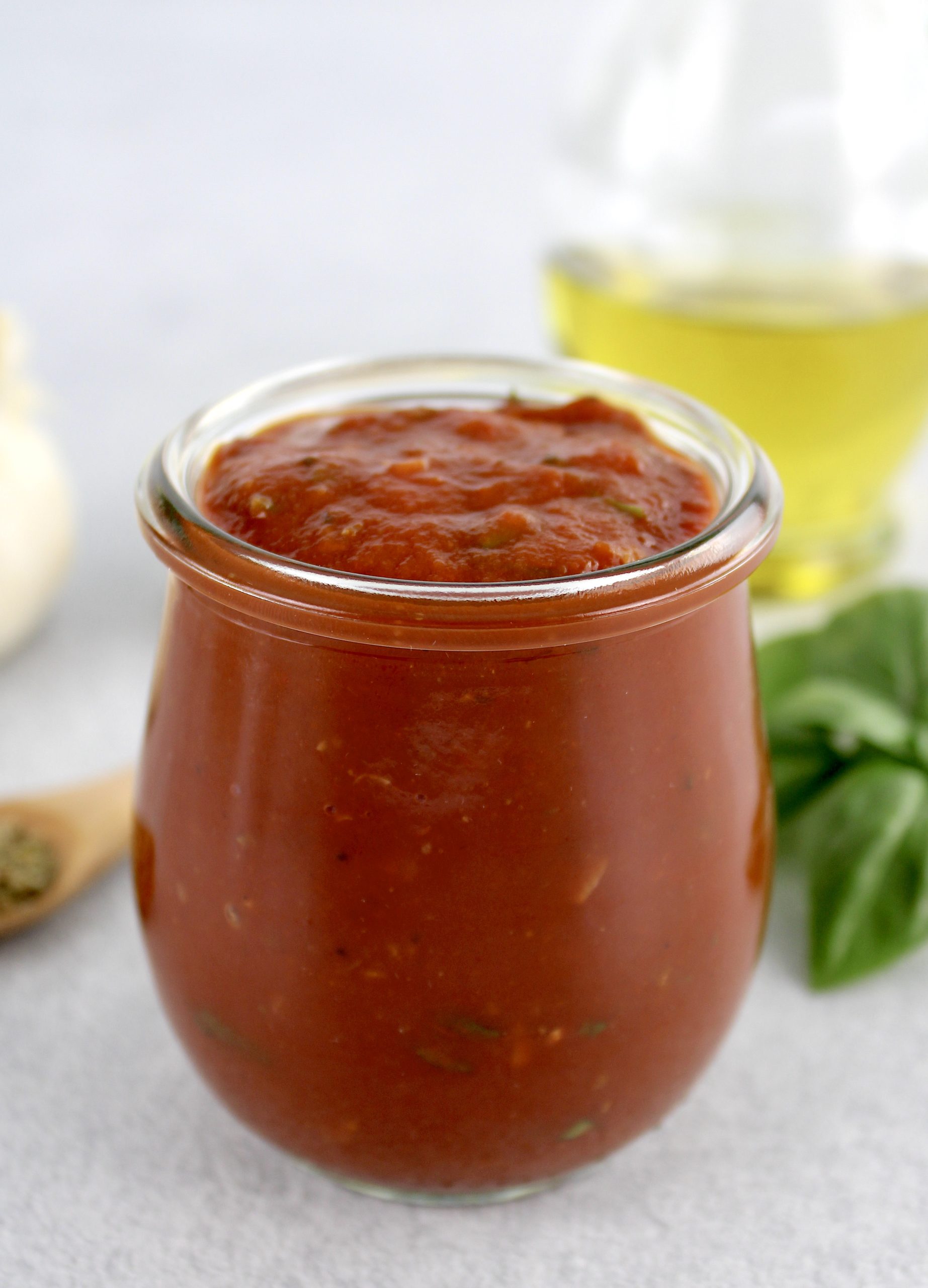 side view of pizza sauce in open glass jar with basil and olive oil in background