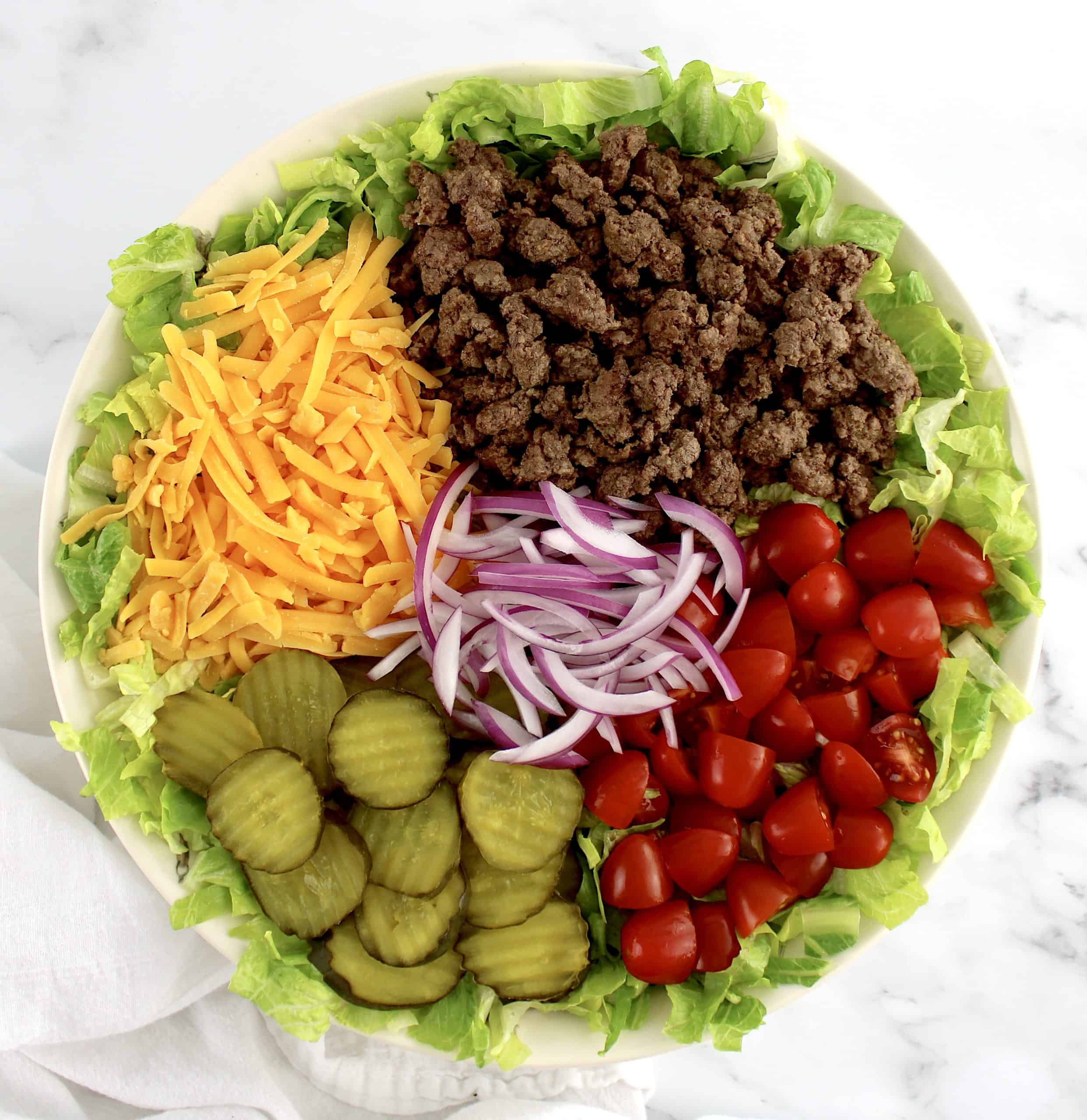 salad in bowl with cooked ground beef pickles tomatoes shredded cheddar and sliced red onions