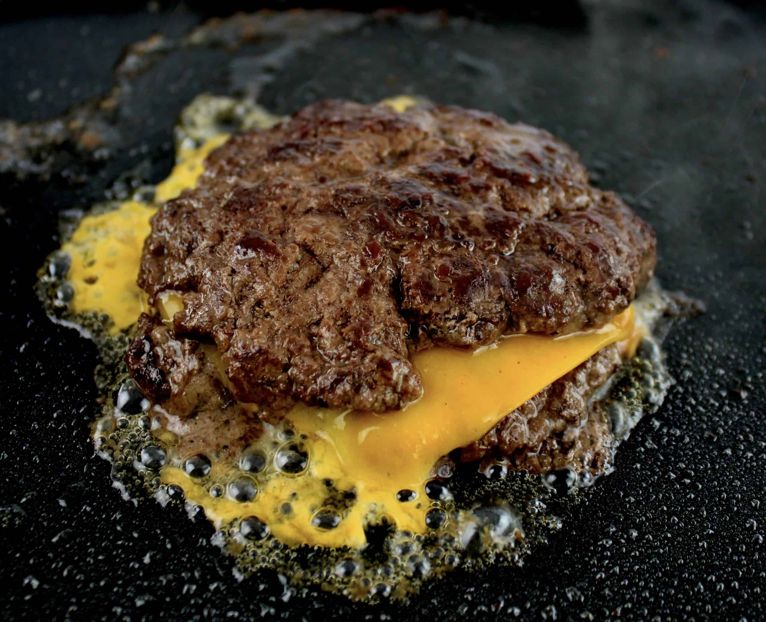 2 burger patties cooking on griddle with slice of melted cheddar cheese in between
