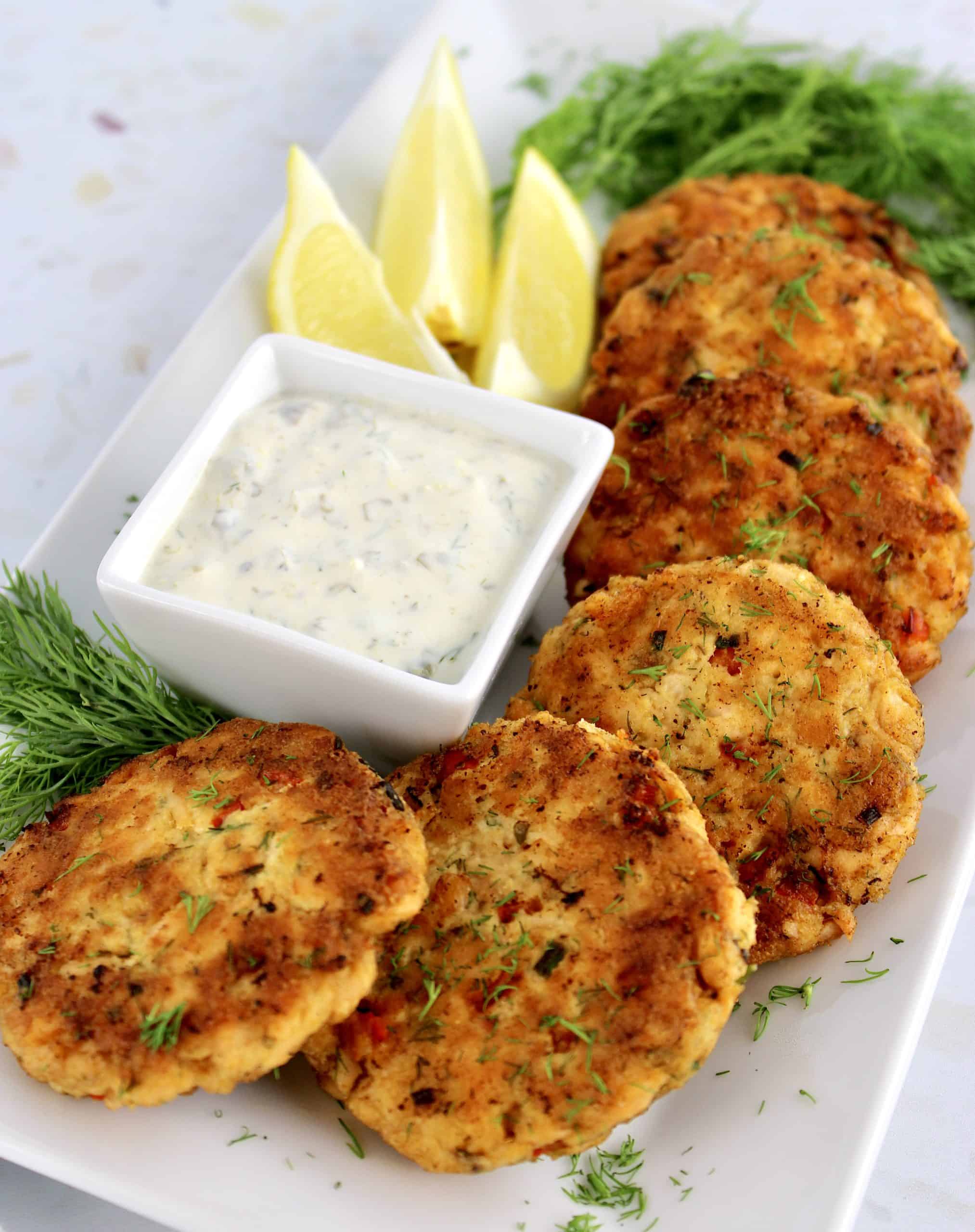 salmon patties on white plate with square dish of dill sauce lemon wedges and fresh dill on the side