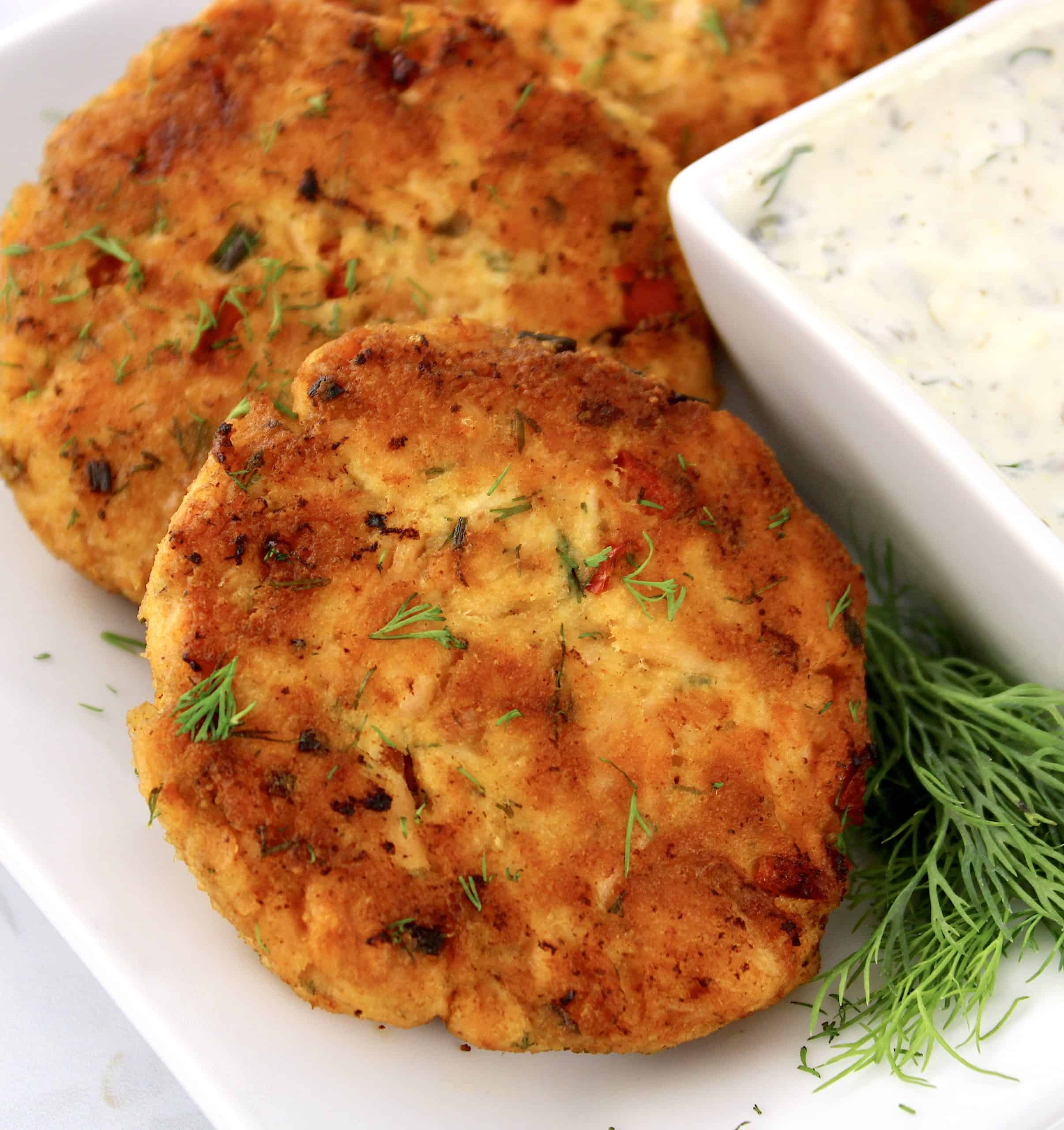 closeup of salmon patties on white plate with fresh dill and dill sauce on side