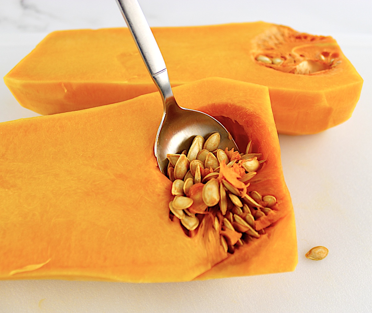 spoon scooping out the seeds of butternut squash on white cutting board