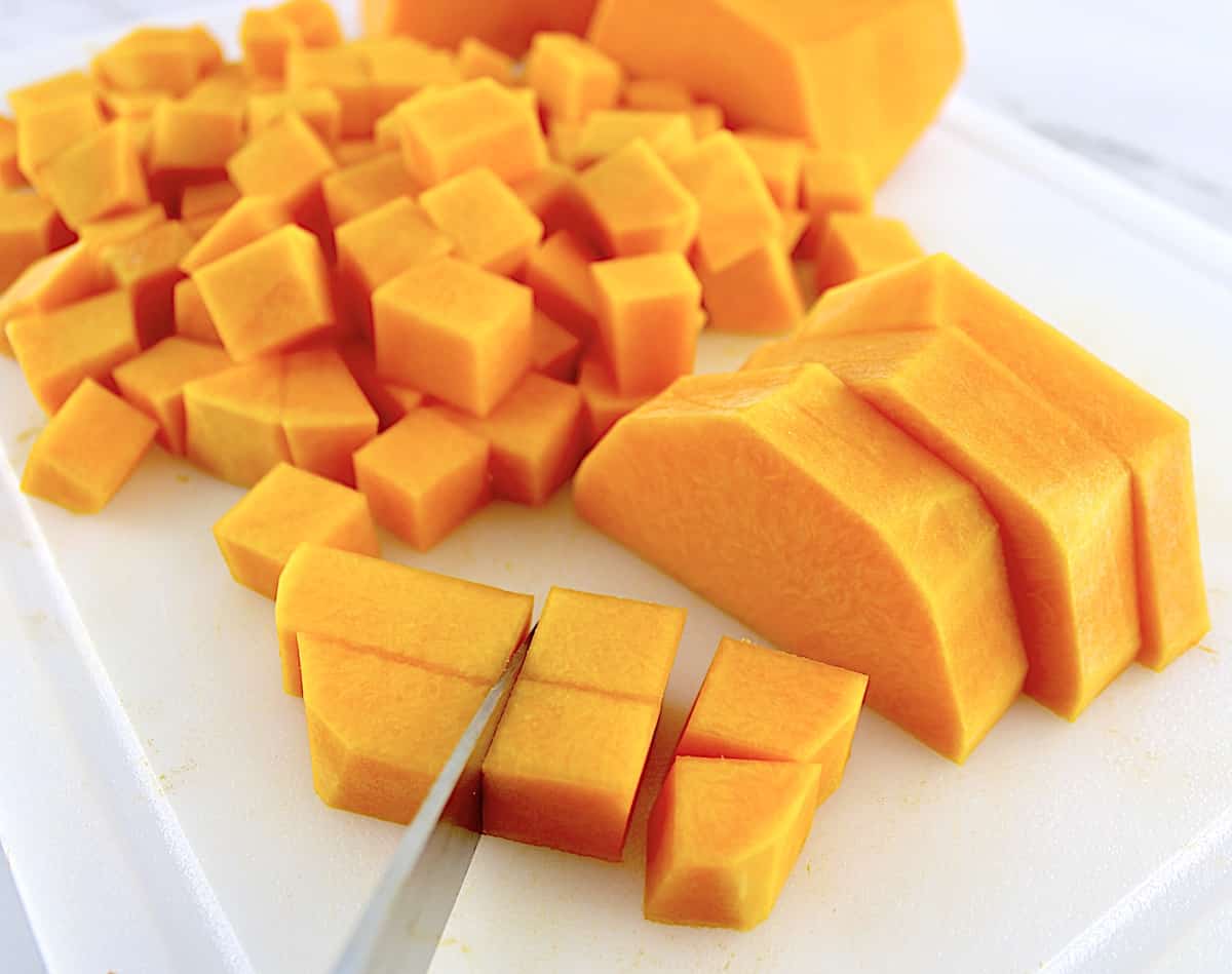 butternut squash being cut into cubes on white cutting board