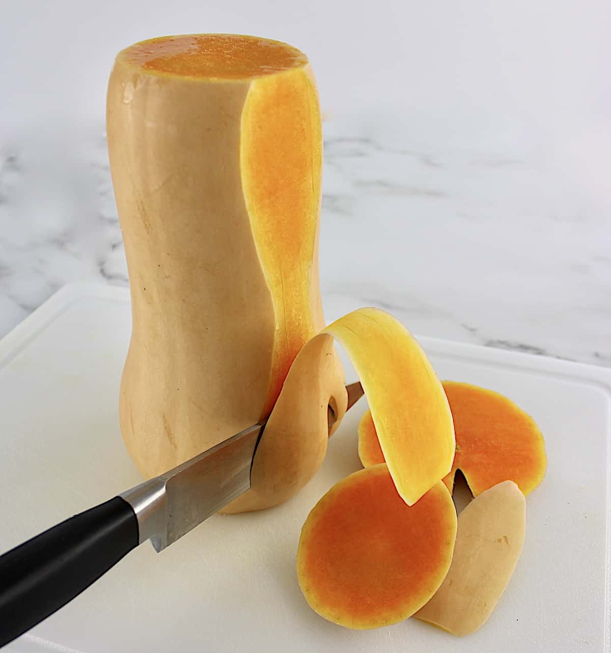 butternut squash being peeled on white cutting board with knife slicing downward