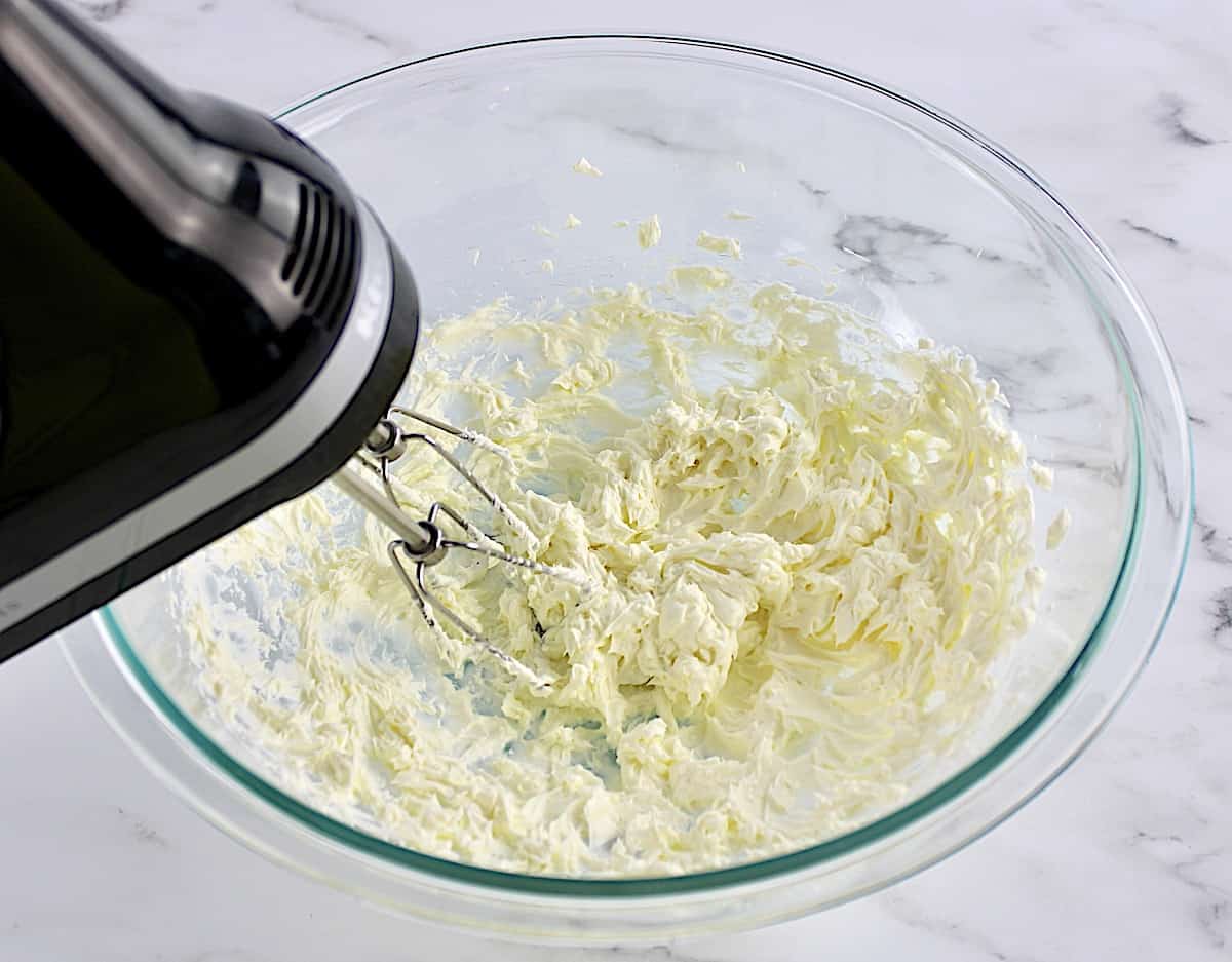 cream cheese being whipped with hand mixer in glass bowl