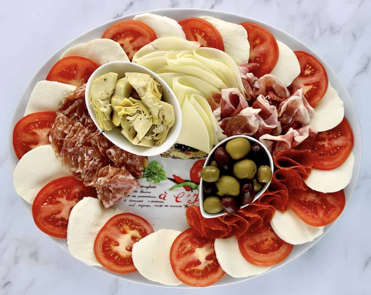 slices of mozzarella cheese and tomatoes around the edge of a platter with olives cheese,cold cuts and artichoke hearts in the center