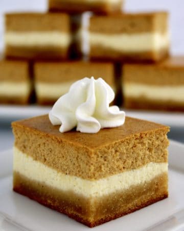 closeup of Keto Pumpkin Cheesecake Bar on white plate with whip cream on top and more bars stacked up in background