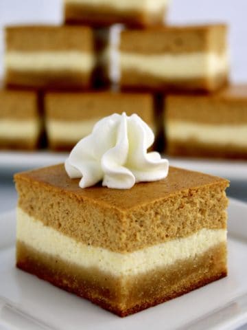 closeup of Keto Pumpkin Cheesecake Bar on white plate with whip cream on top and more bars stacked up in background