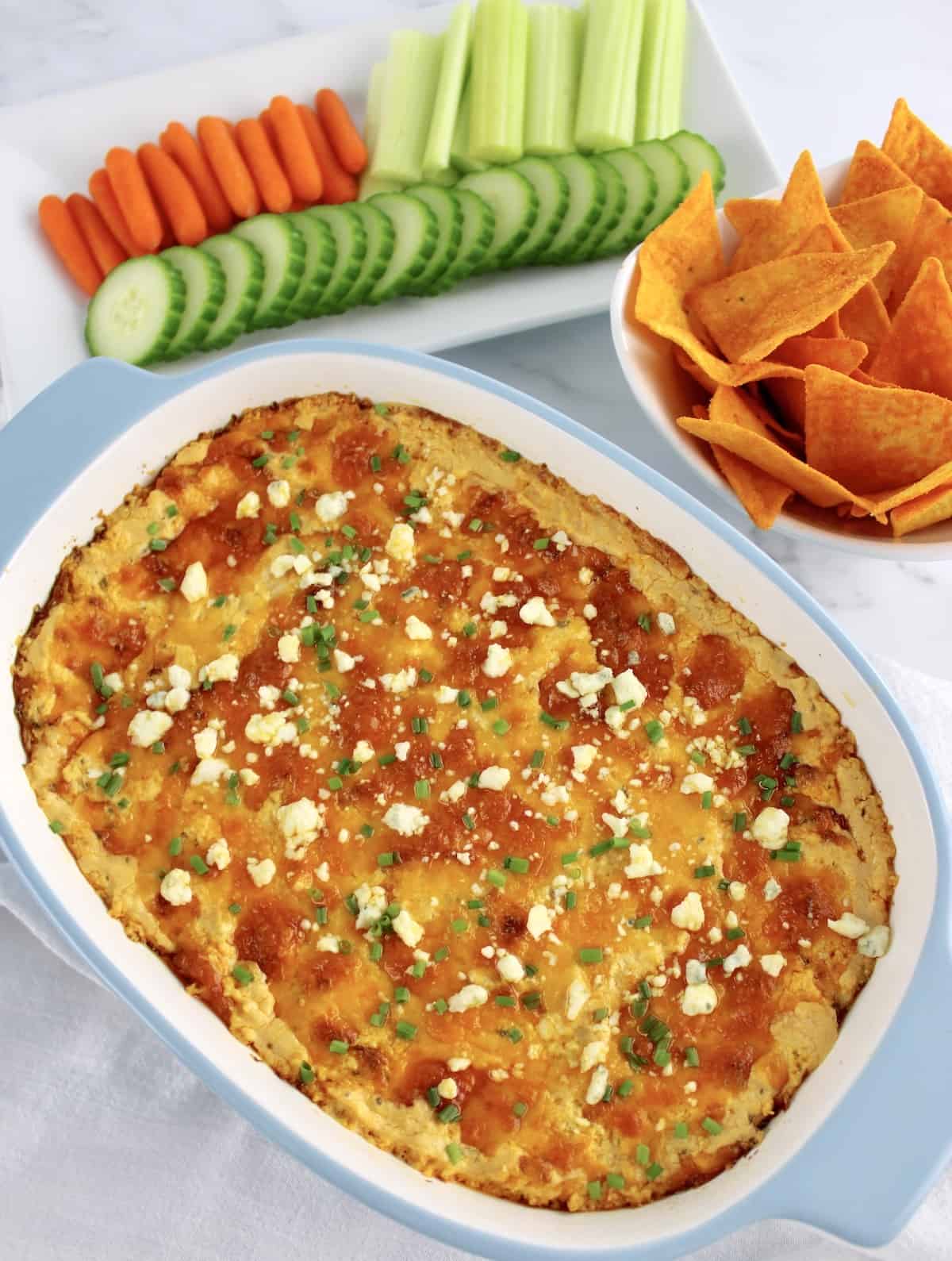 Buffalo Chicken Dip in baking dish with sliced veggies and tortilla chips in background