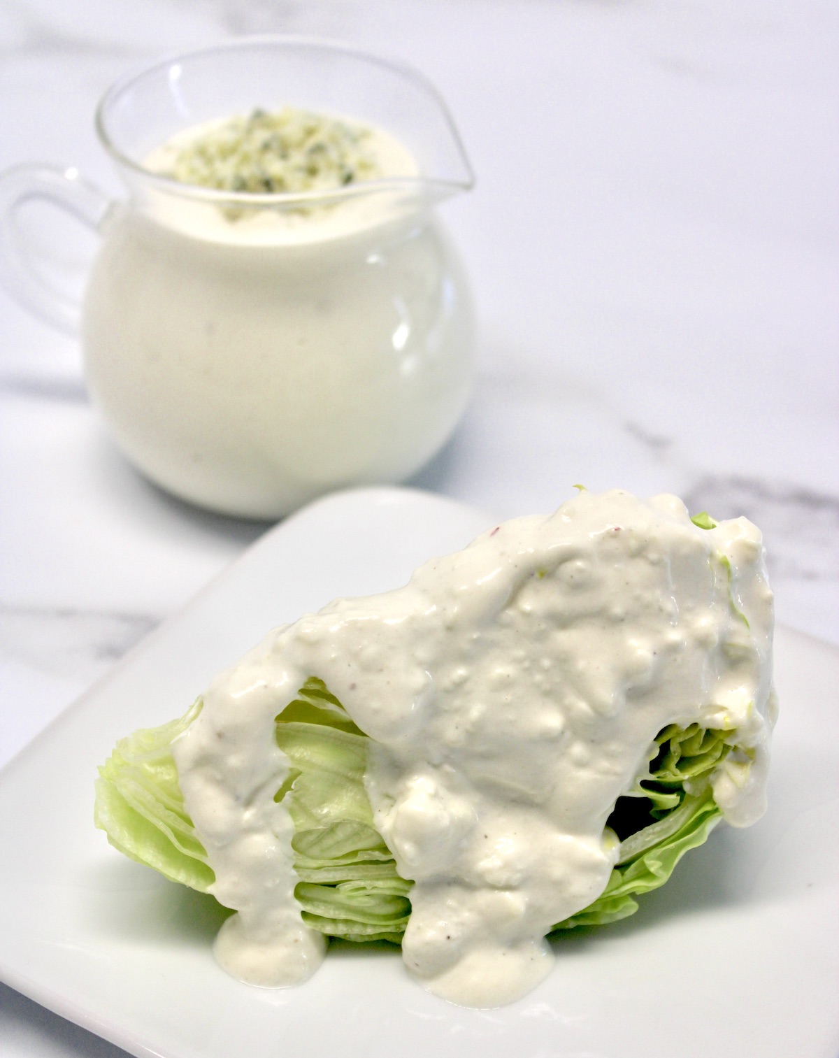 Keto Blue Cheese Dressing over iceberg wedge on white plate with glass pitcher in background