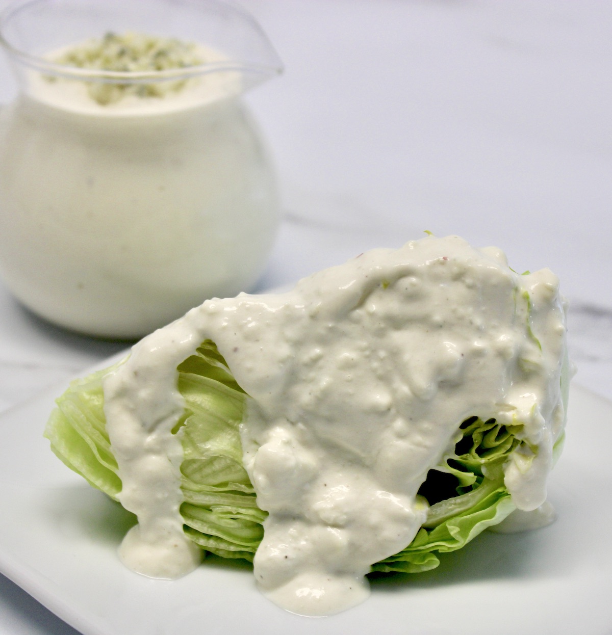 Keto Blue Cheese Dressing over iceberg wedge on white plate with glass pitcher in background