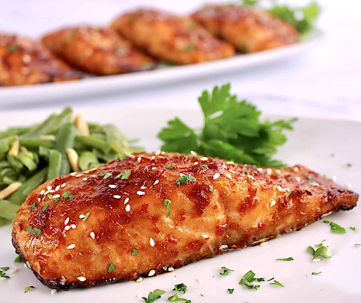 closeup of Asian Glazed Salmon on white plate with parsley and more salmon in background