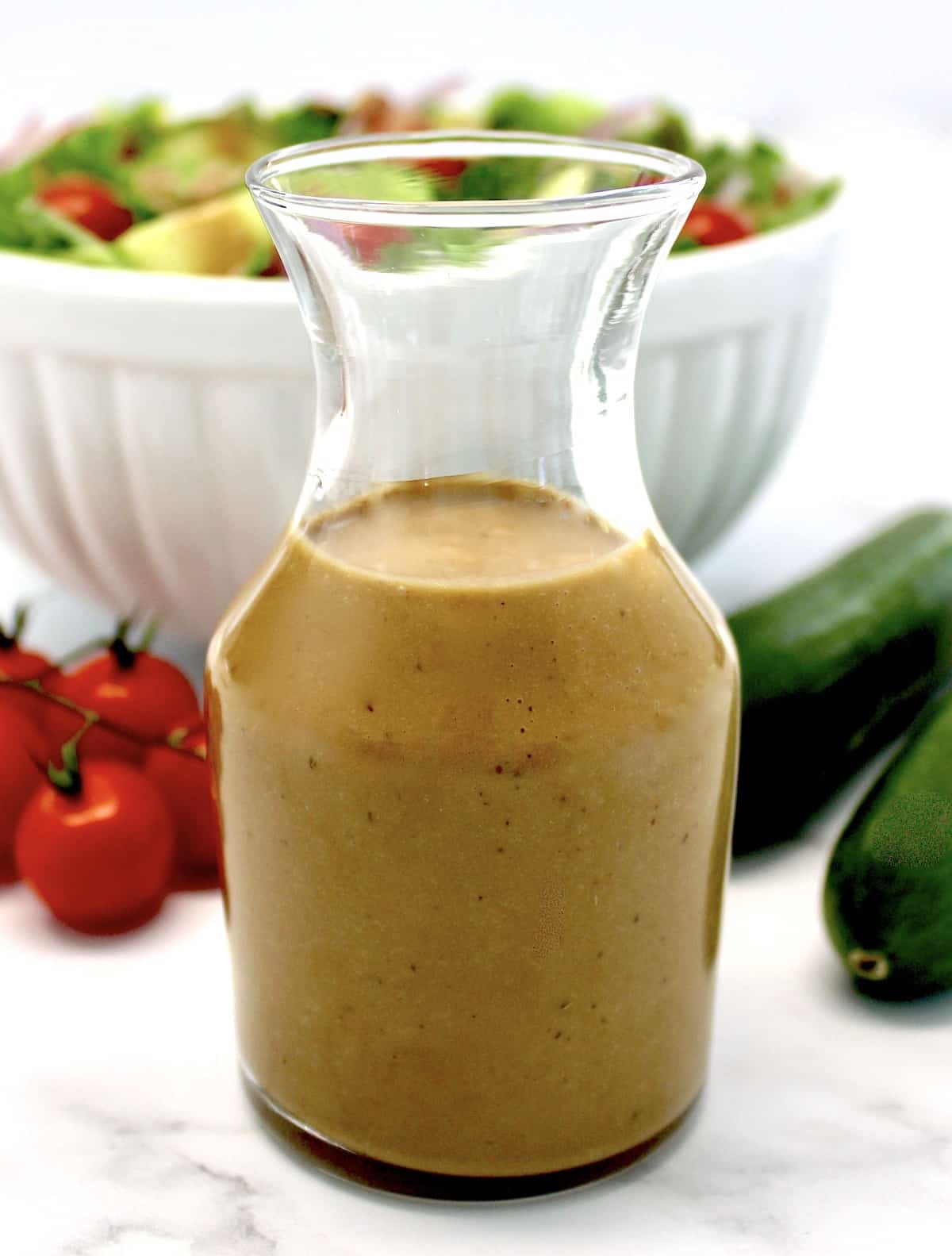 closeup of Creamy Balsamic Dressing in glass container with salad, tomatoes and cucumbers in background