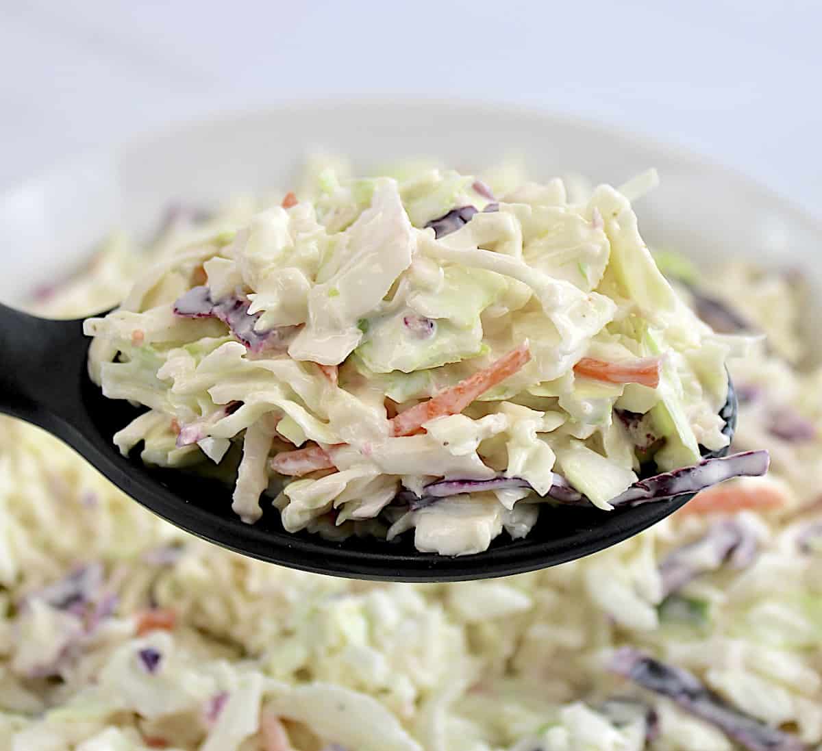 Easy Coleslaw being held up with black serving spoon over bowl of coleslaw