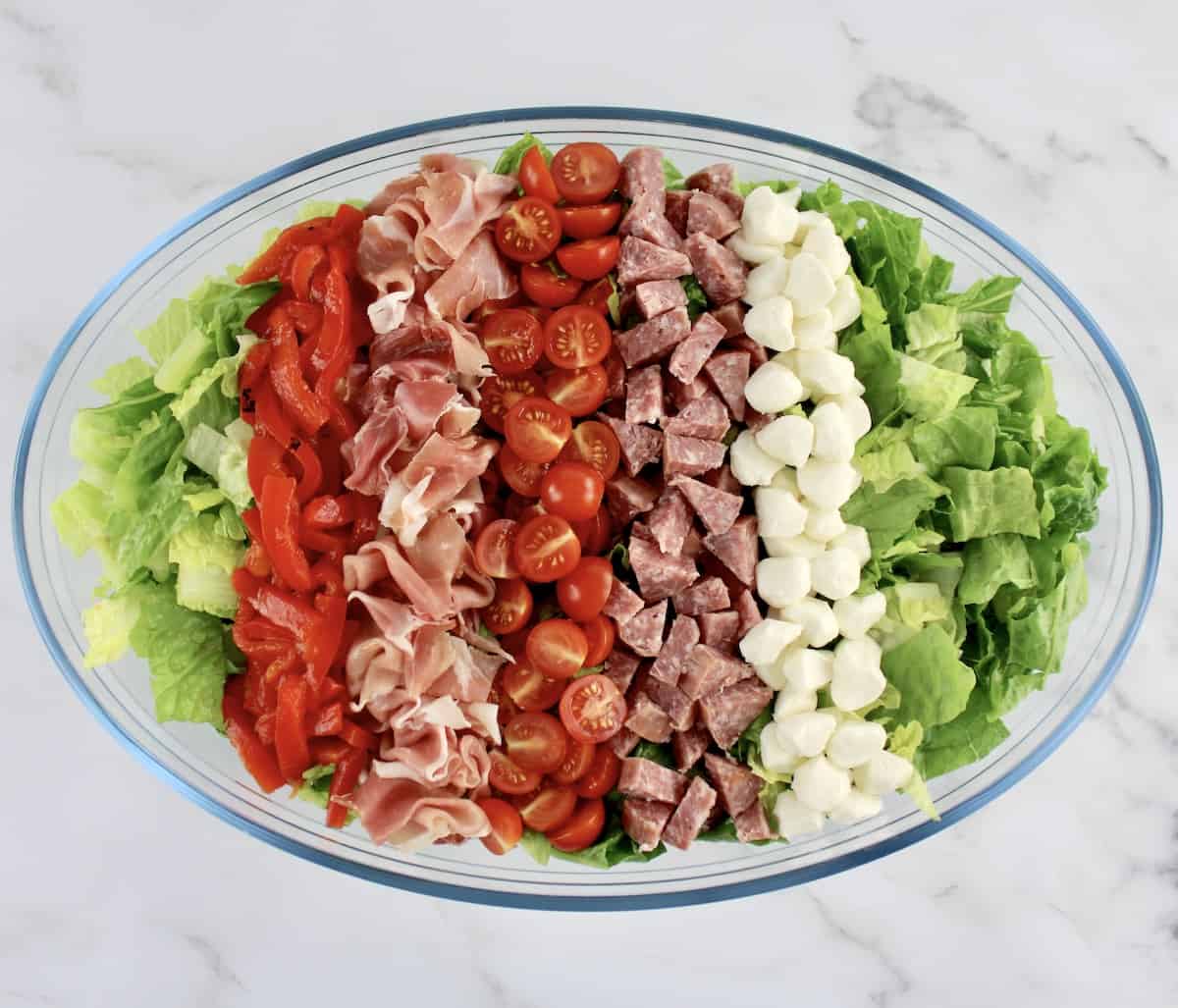 romaine lettuce in oval glass bowl with rows of prosciutto roasted rd pepper strips halved tomatoes mozzarella pearls and chopped soppressata