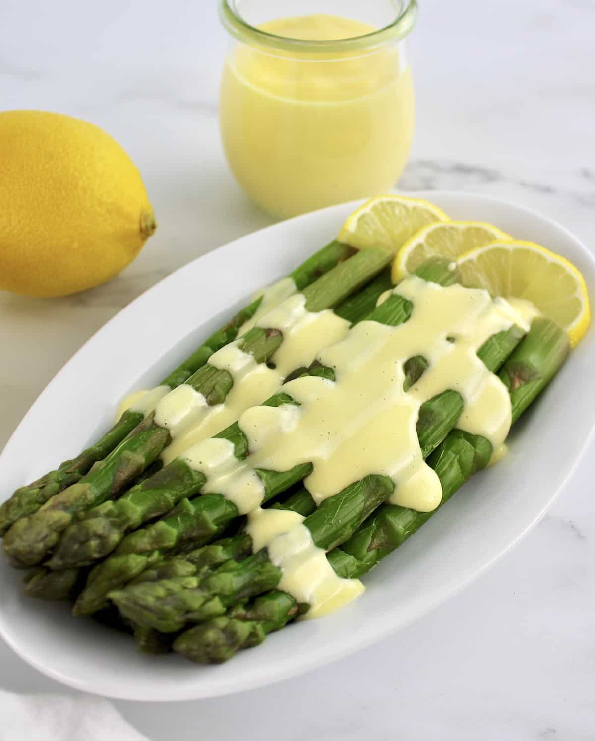 asparagus with Hollandaise Sauce on white plate with more sauce in jar and lemons in background