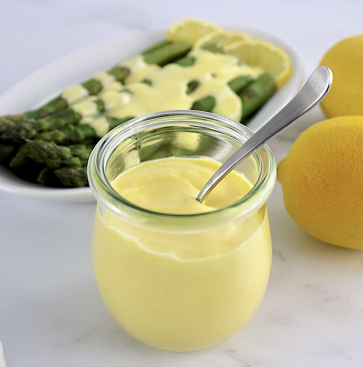 Hollandaise Sauce in open glass jar with spoon in it and asparagus in background