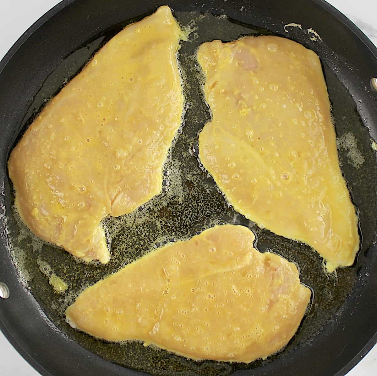 3 chicken cutlets dipped in egg being sauteed in skillet