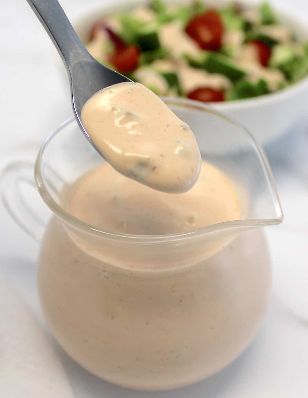 Thousand Island Dressing being spooned out of glass pitcher with salad in background