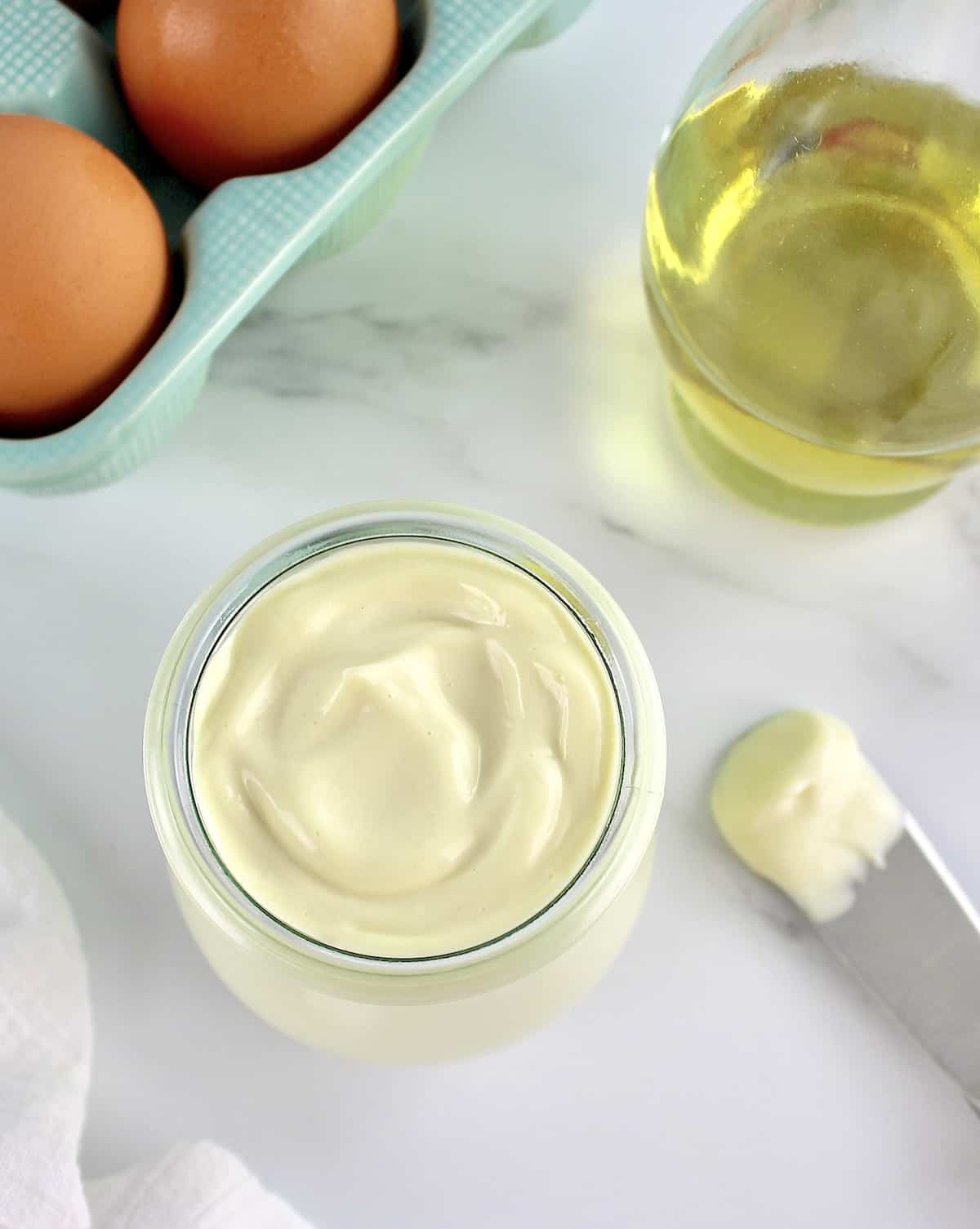 overhead view of Easy Homemade Keto Mayonnaise in open glass jar with eggs and oil in background