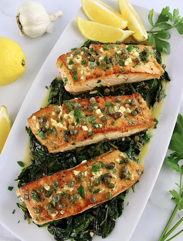 overhead view of 3 pieces of Salmon Piccata with Spinach on white plate with lemon slices in back