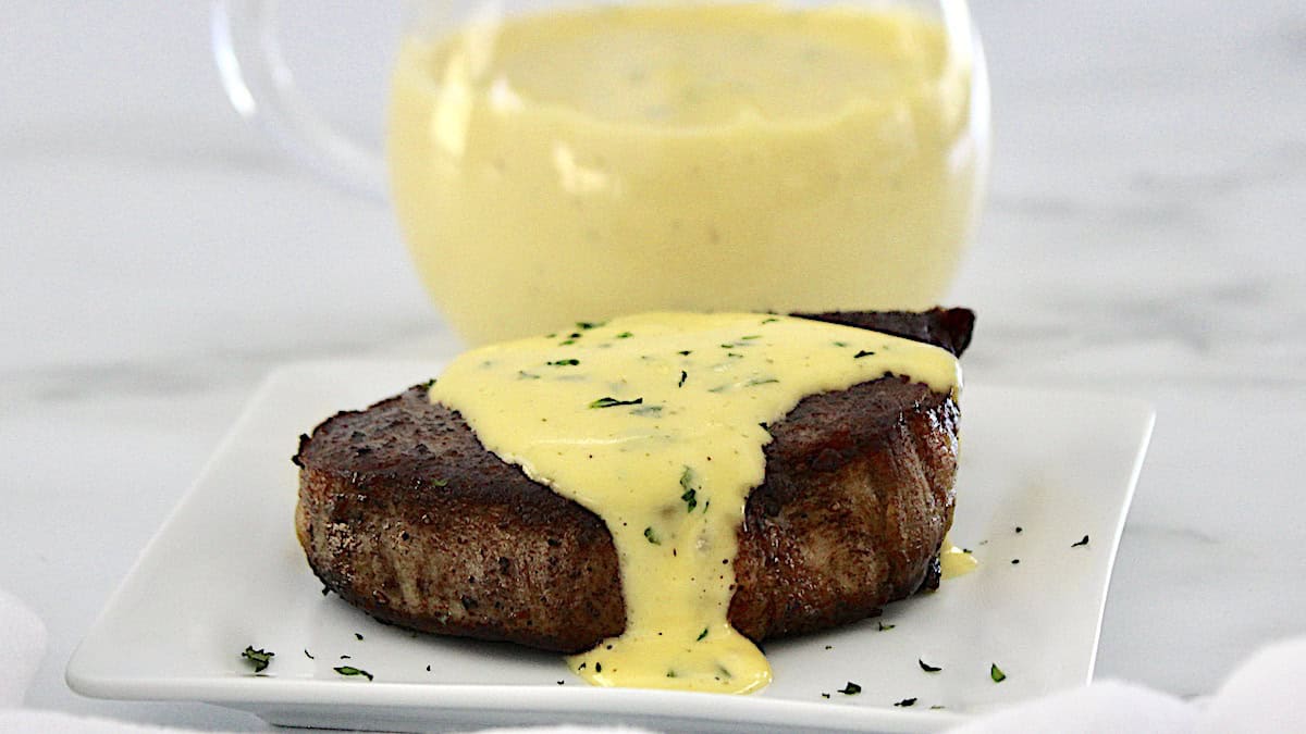 Bearnaise Sauce poured over steak on white plate with more sauce in glass pitcher in back