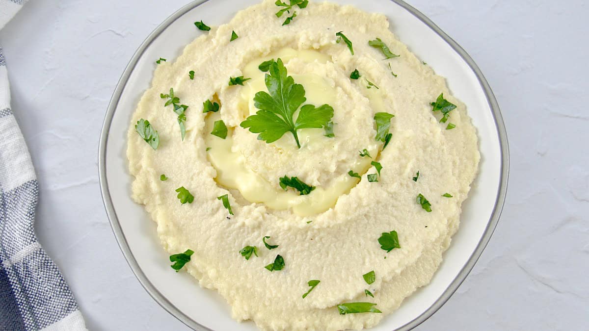 Cheesy Mashed Cauliflower in white bowl with chopped parsley and parsley leaf on top