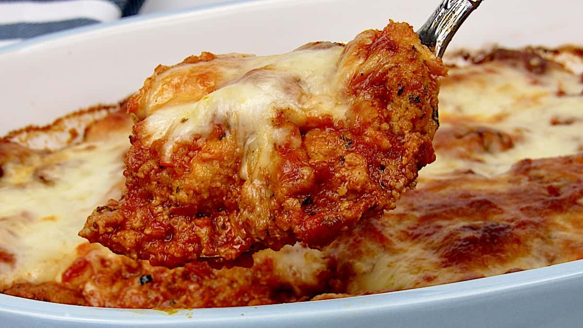 Chicken Parmesan Casserole in silver serving spoon lifting out of blue casserole