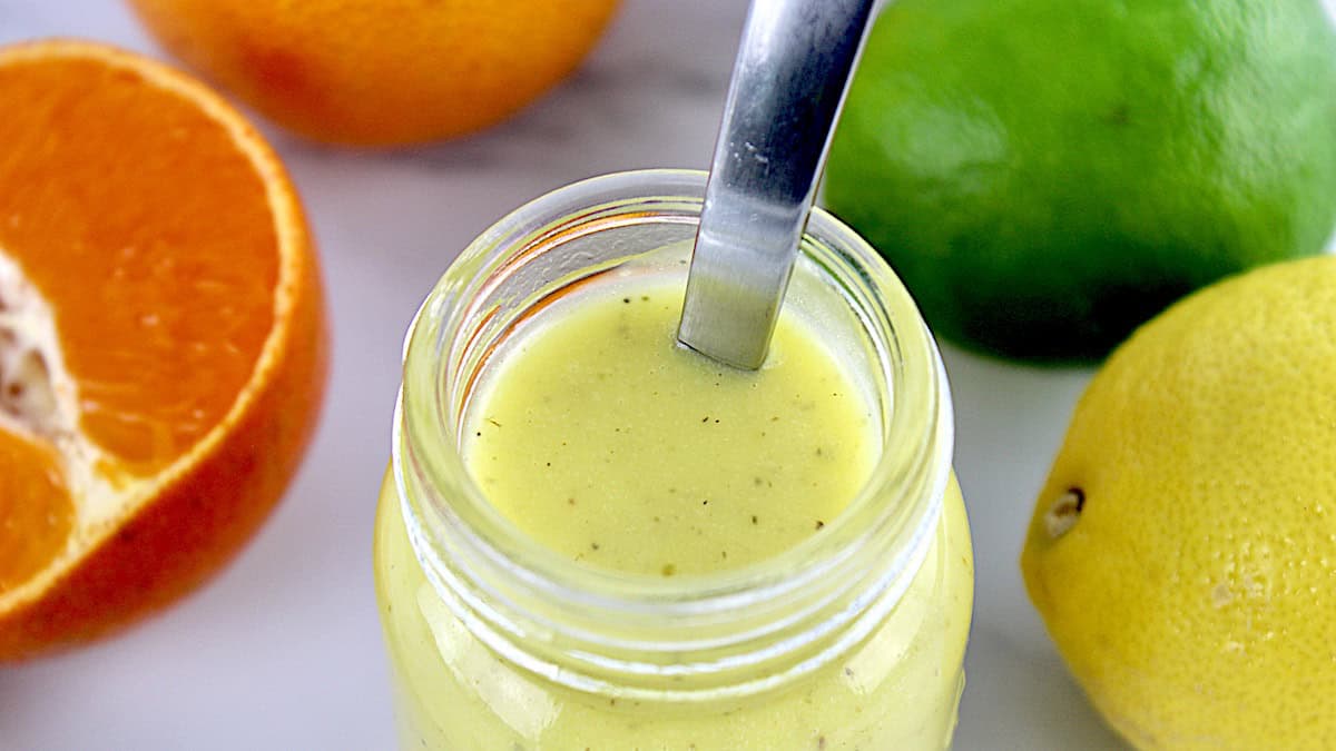 overhead view of Citrus Vinaigrette in open glass jar with spoon in it and oranges and lime in background