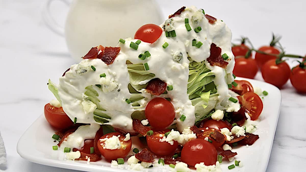 closeup of Classic Wedge Salad with blue cheese dressing dripping from the top and bacon, chives and slices tomatoes